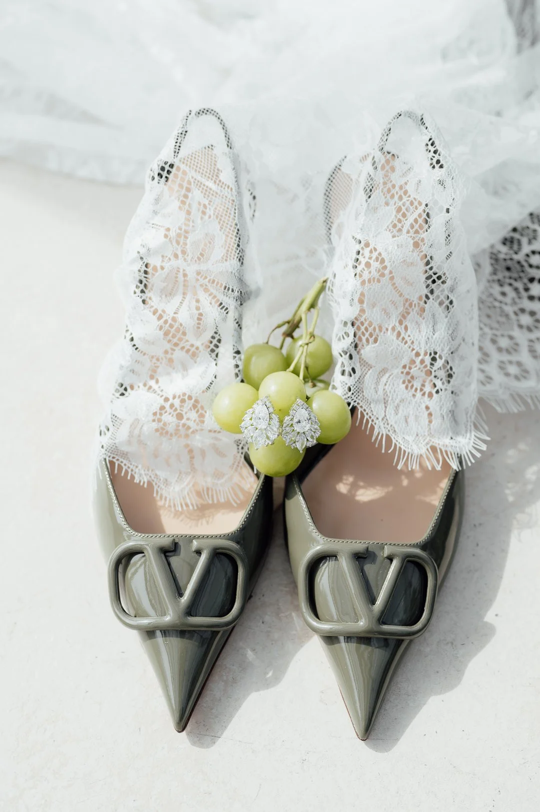 Pair of pointed-toe Valentino high heels with black, white, and gray color blocking, featuring large gray 'V' logos on the toes, lace tights with floral patterns, a bunch of green grapes, and chandelier earrings with large white stones.