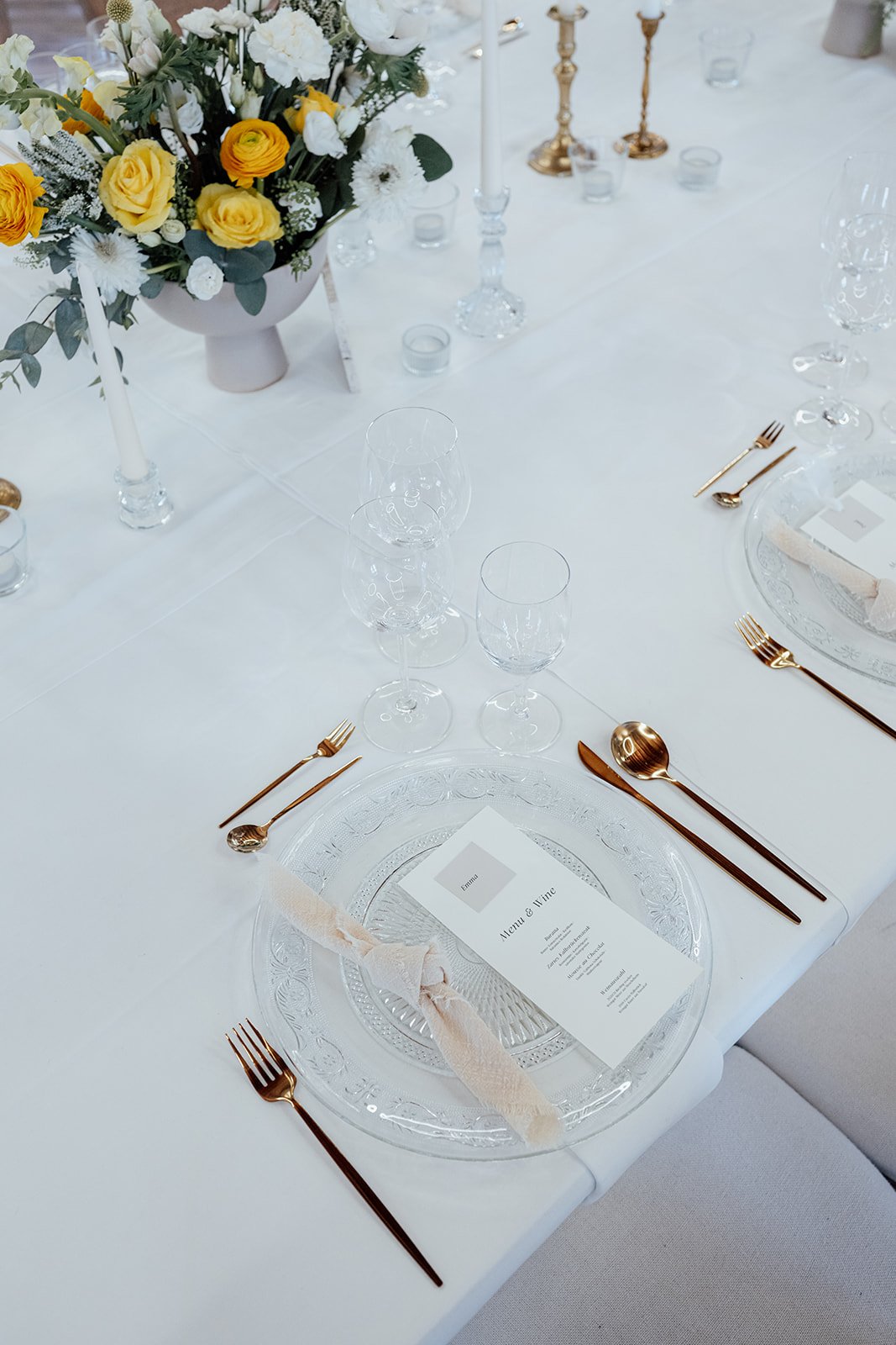 Bild eines gedeckten Festtisches. Auf dem gläsernen Platzteller ist eine helle Stoffserviette und eine Menükarte. Neben dem Teller ist goldenes glänzendes Besteck. Außerdem stehen viele Weingläser auf dem Tisch.
