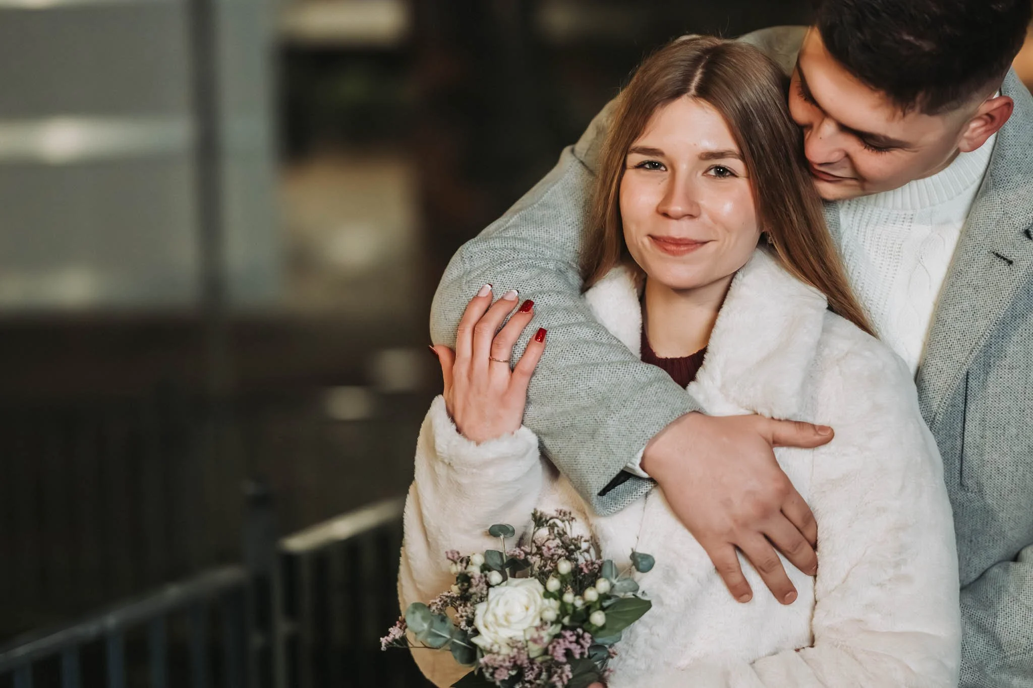 Portrait von einer Frau, die lächelnd in die Kamera sieht. Sie wird von hinten von einem Mann umarmt, der lächelt. Sie hat einen kleinen Blumenstrauß vor sich und hält seinen Arm mit einer Hand fest. Beide tragen Mäntel und es ist nacht.