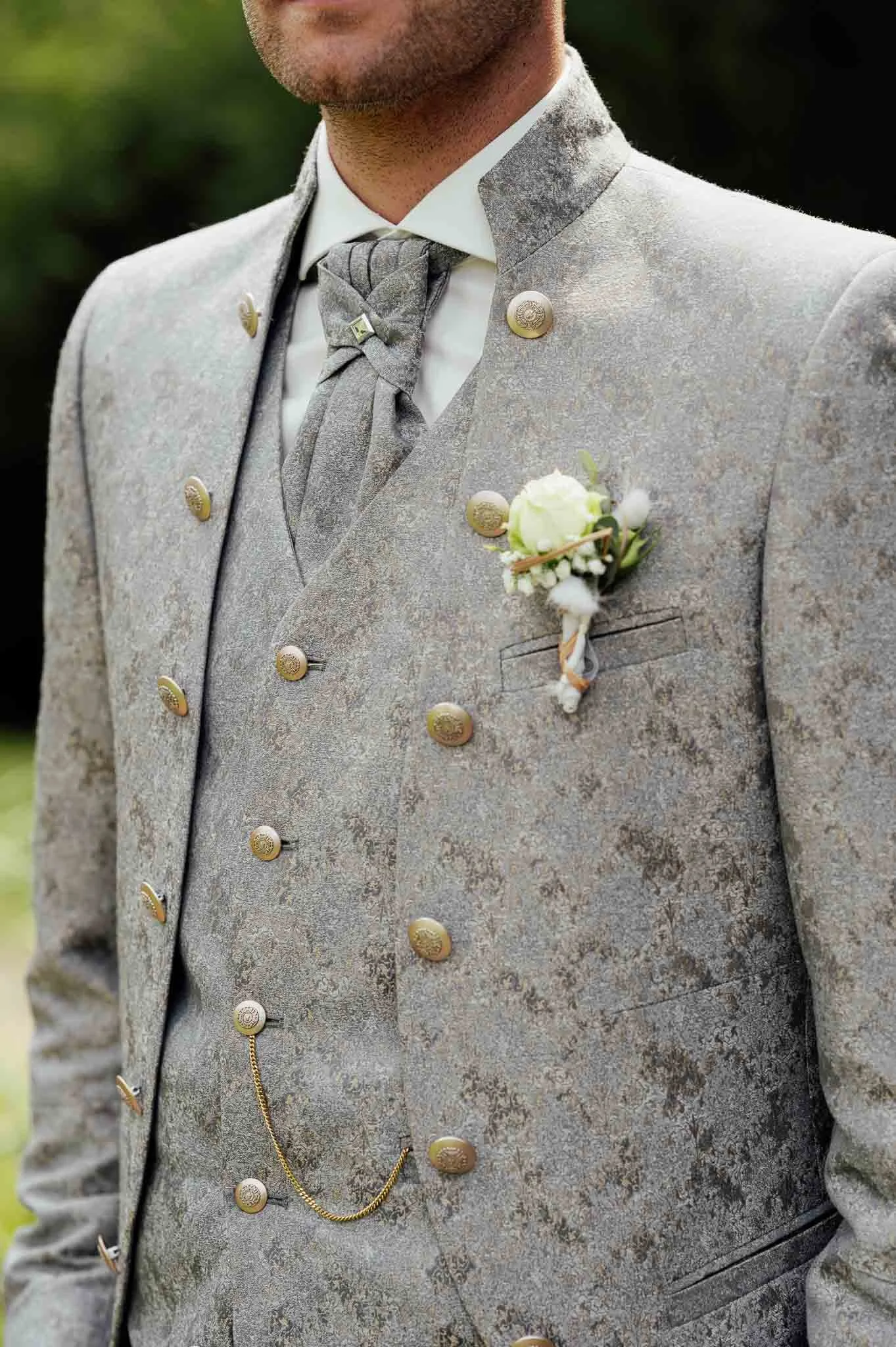 Nahaufnahme eines Mannes in traditioneller, eleganter Trachtenjacke mit Blumen-Anstecker, Krawatte und Damenkette, vermutlich bei einer Feier oder Hochzeit.