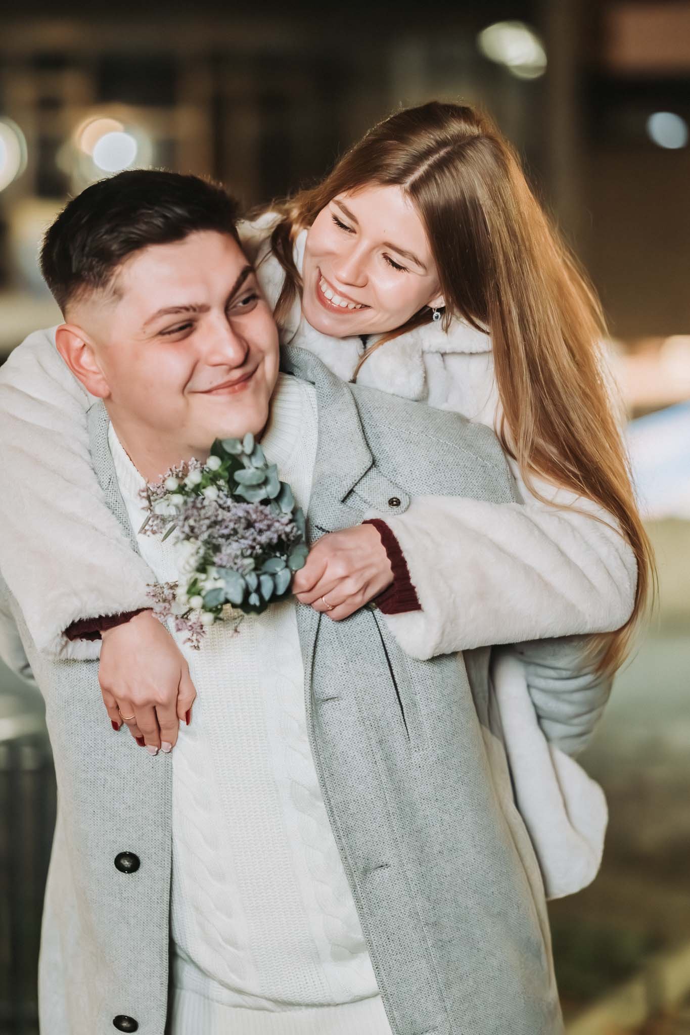 Ein Mann trägt eine Frau auf dem Rücken, beide lachen und sind winterlich angezogen. Die Frau hat einen kleinen Blumenstrauß in der Hand