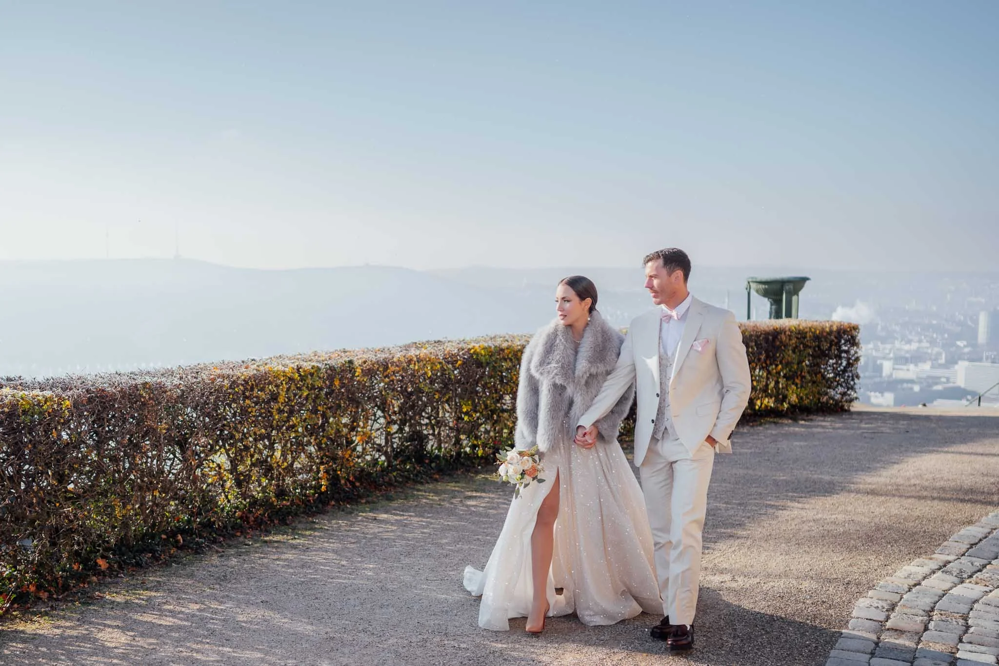 Ein frisch vermähltes Paar beim Spaziergang auf einer Stadtterrasse mit Sicht auf die Stadt und die Berge im Hintergrund. Die Frau trägt ein schimmerndes Brautkleid und einen grauen Pelz, hält einen Brautstrauß, der Mann trägt einen weißen Anzug.
