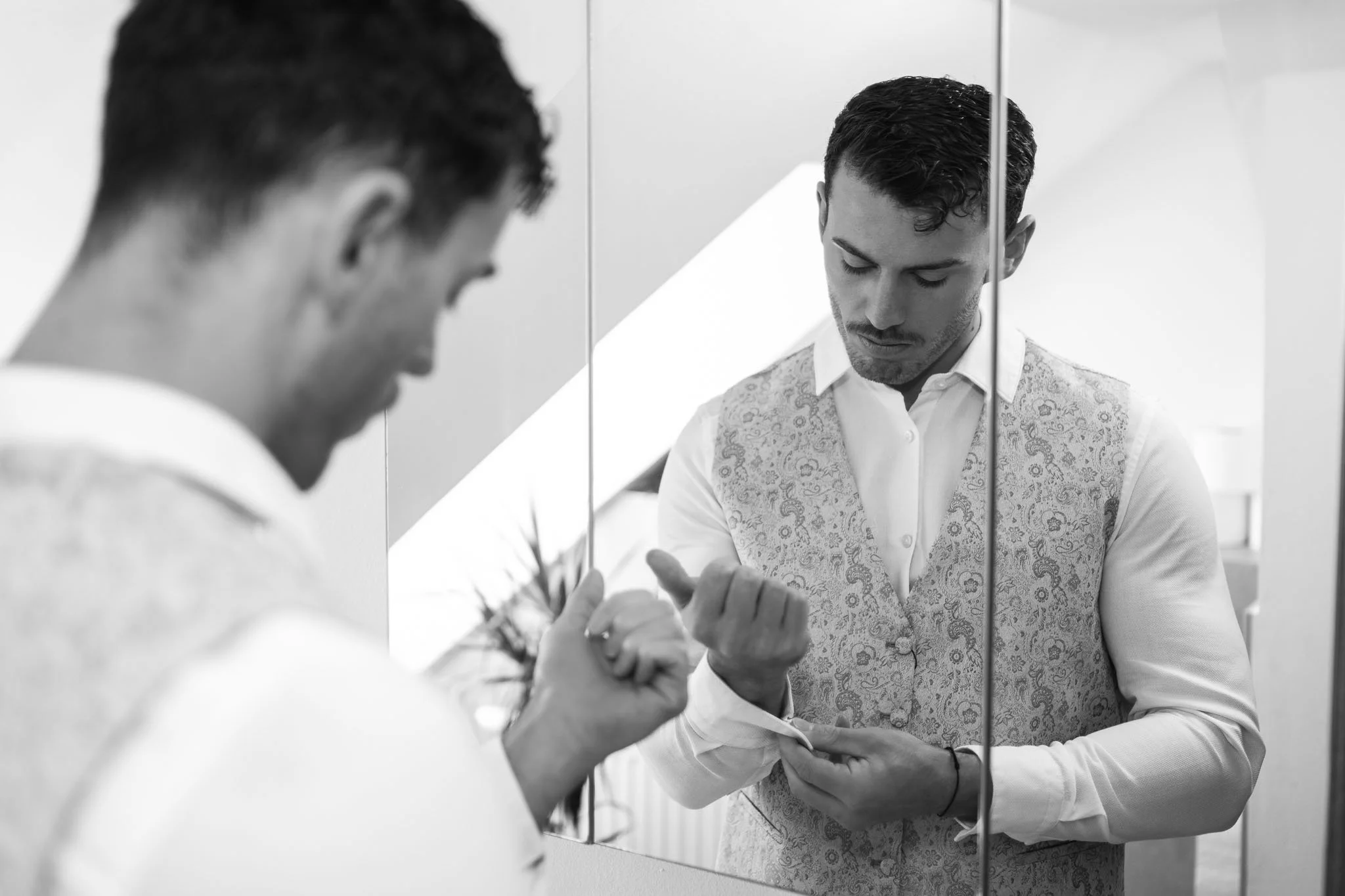 Junger Mann steht vor einem Spiegel, trägt ein weißes Hemd und eine Weste, und schaut auf seine aufgeblasene Hand. Die Szene ist in Schwarz-Weiß gehalten.