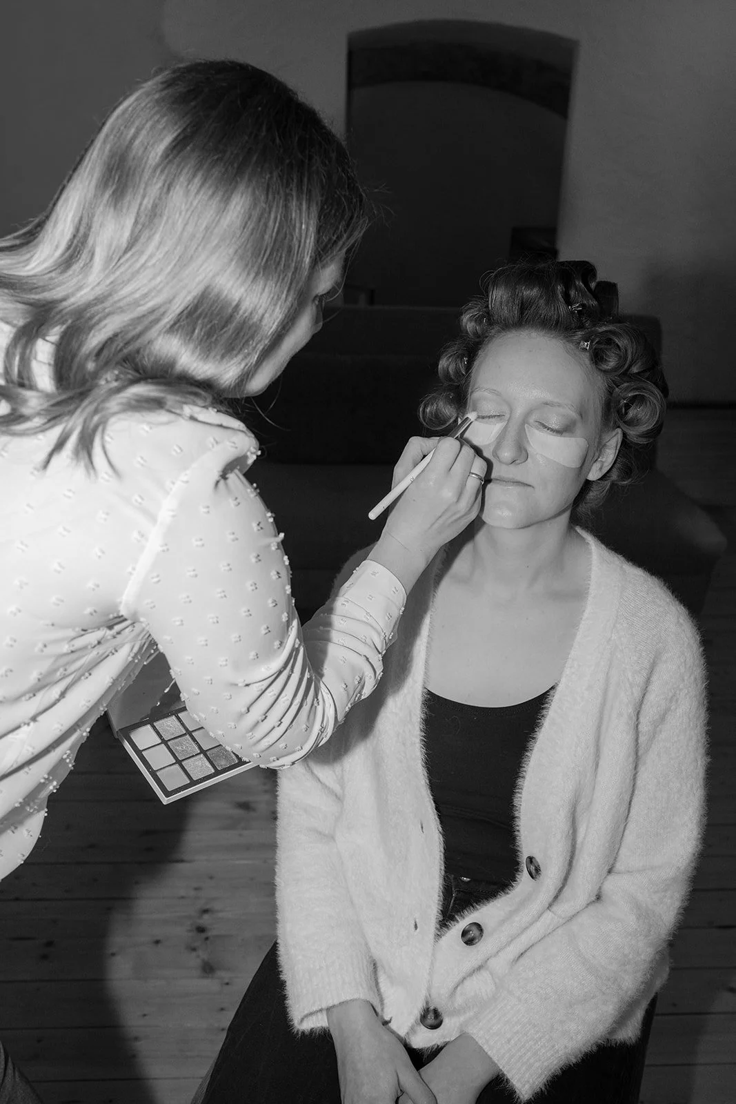Schwarzweiß Bild von einer Frau, die Lockenwickler auf dem Kopf hat und von einer anderen Frau geschminkt wird.