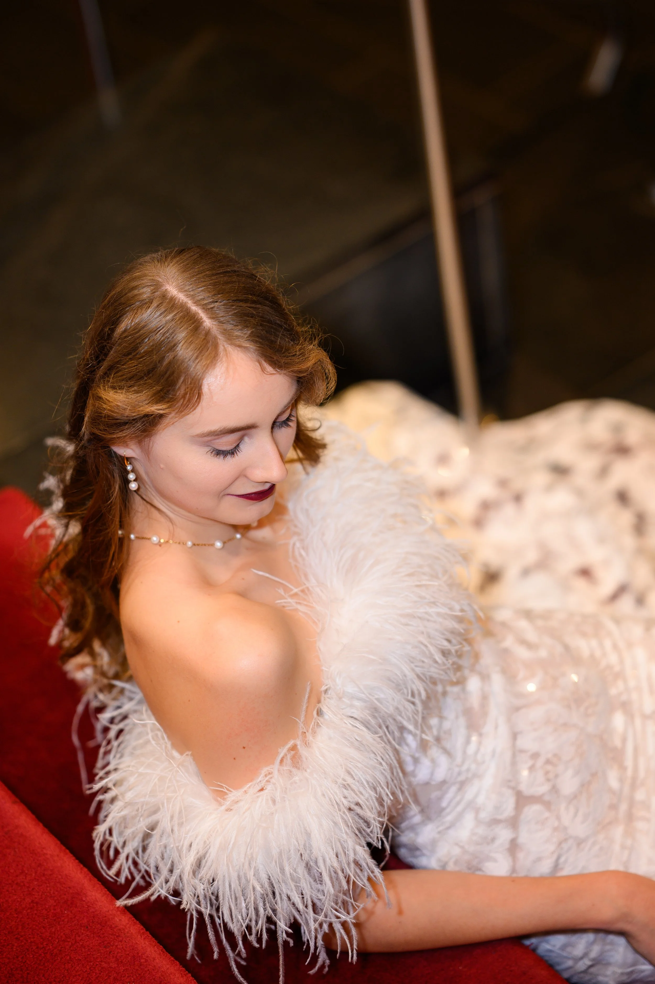 Bild von leicht schräg oben einer Braut, die auf einer Treppe sitzt und sich elegant auf ihre nackte Schulter schaut. Sie trägt ein extravagantes Kleid mit Federn und Perlenschmuck