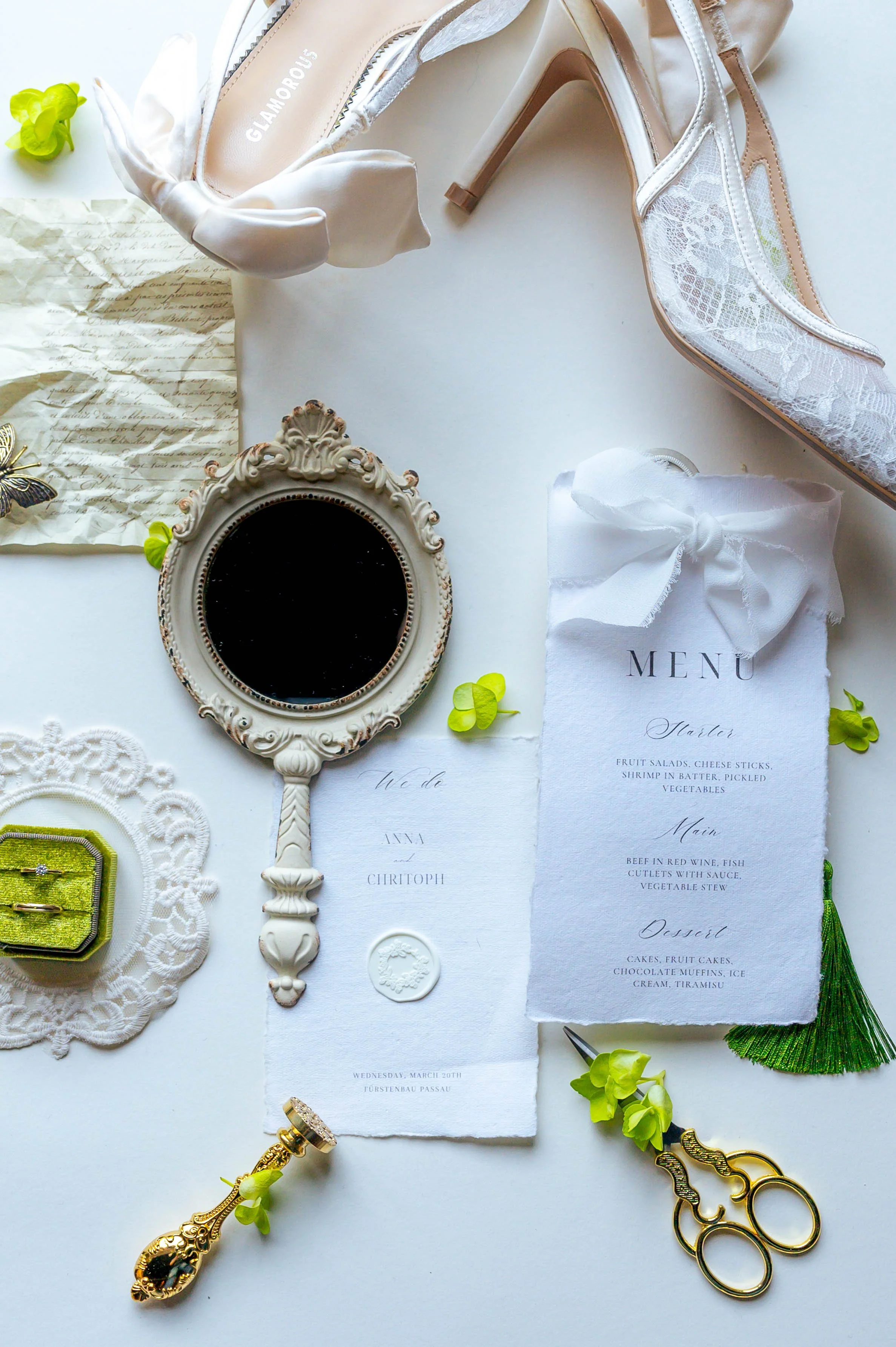 Hochzeitsdetails mit Schuhen, Fernbedienung, Brief, Menükarte, Schmuck, Schneidewerkzeug, grünen Blumen und Dekorationsgegenständen auf weißem Hintergrund.