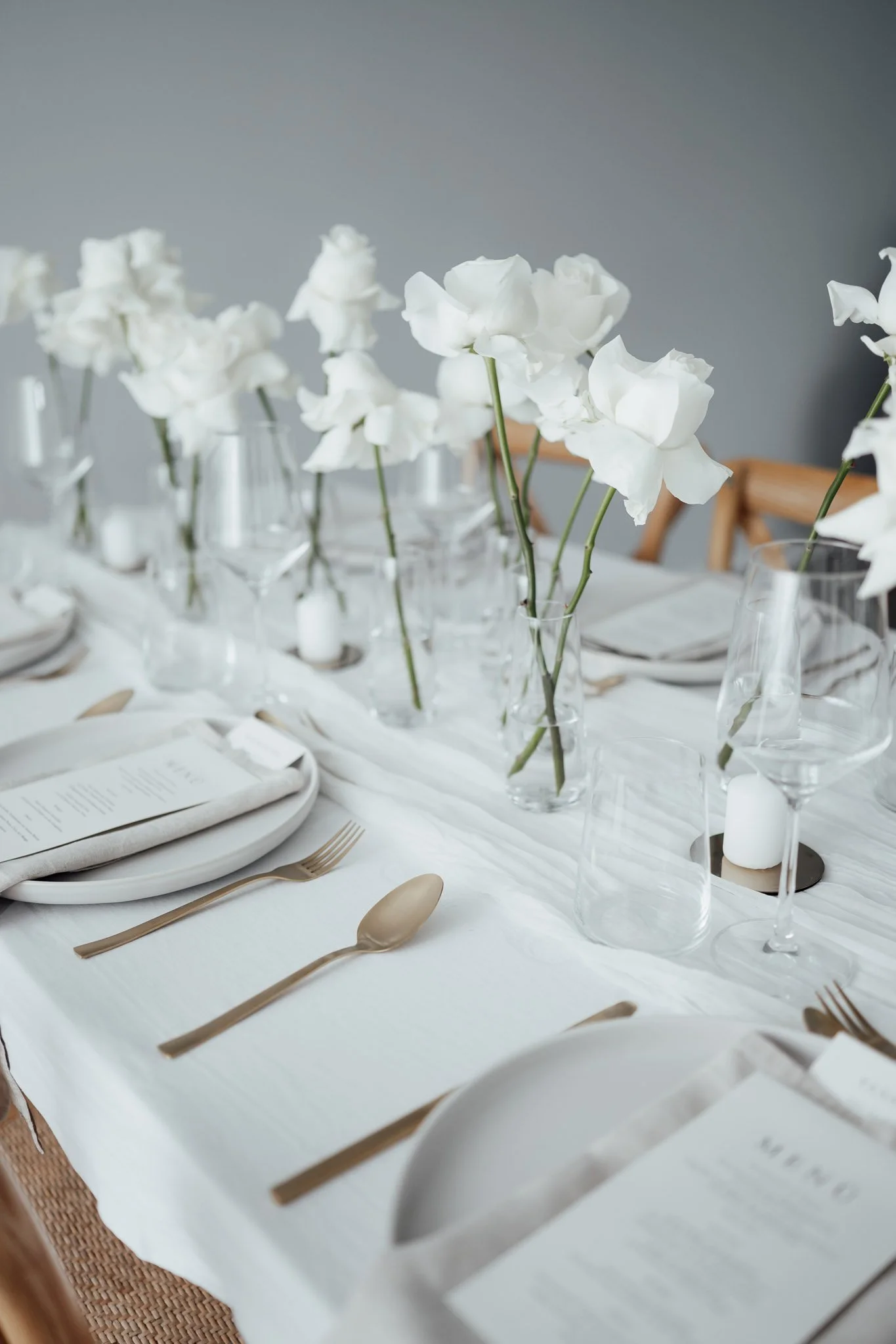 Ein eleganter Esstisch mit weißen Blumen in Gläsern, Besteck, Tellern, Weingläsern und Kerzen auf einer weißen Tischdecke.