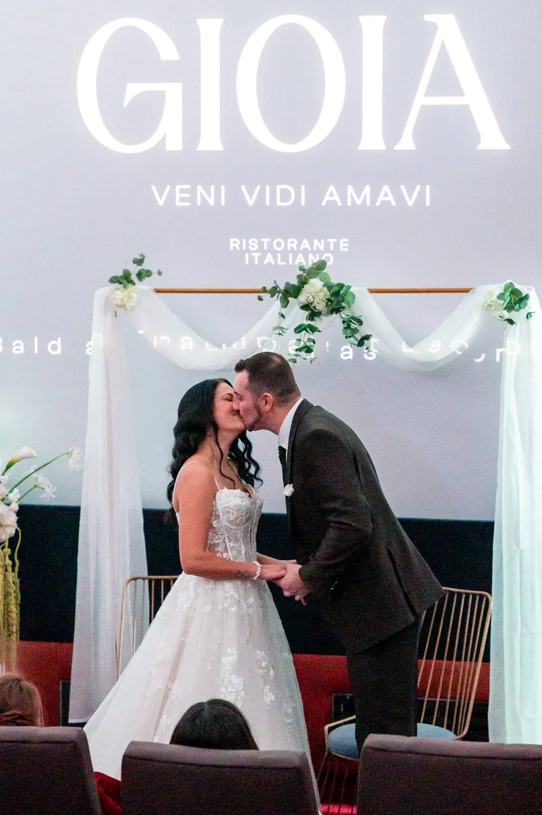 Ein Brautpaar küsst sich vor einer Leinwand im Kinosaal, vor der ein goldener Traubogen mit weißem Stoff und Blumen steht