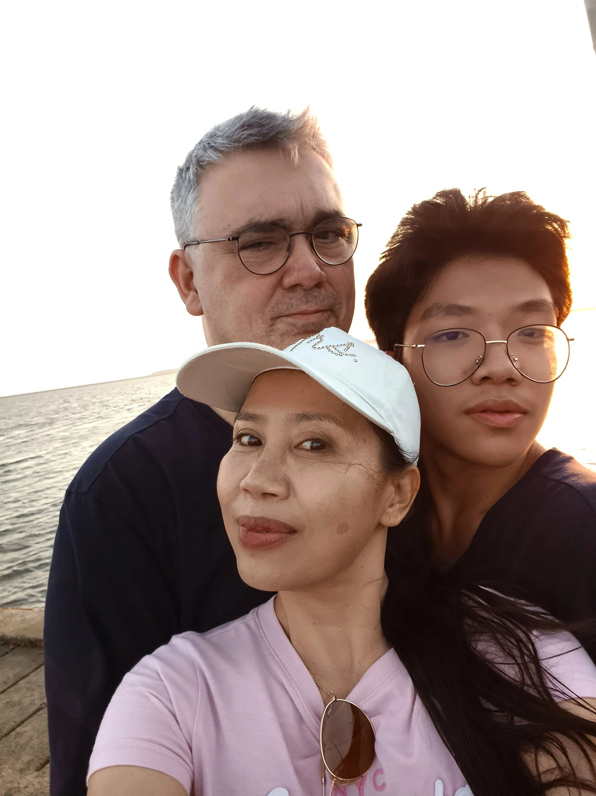 Vier Menschen, drei Erwachsene und ein Jugendlicher, stehen am Wasser bei Sonnenuntergang und machen ein Selfie. Sie tragen Brillen und sommerliche Kleidung, das Mädchen hat eine weiße Kappe auf. Im Hintergrund sieht man das Meer und den Himmel bei Sonnenuntergang.