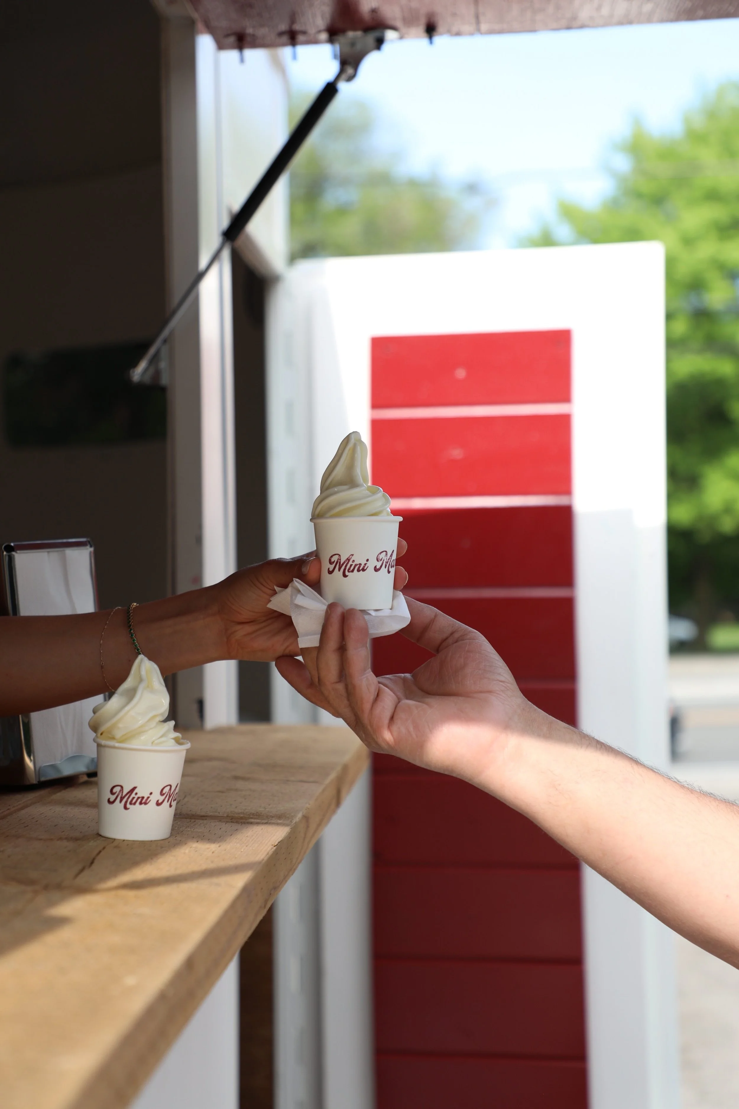 A person is handing a soft serve ice cream cone to another person at an outdoor stand, with another ice cream on the counter and a red and white background.
