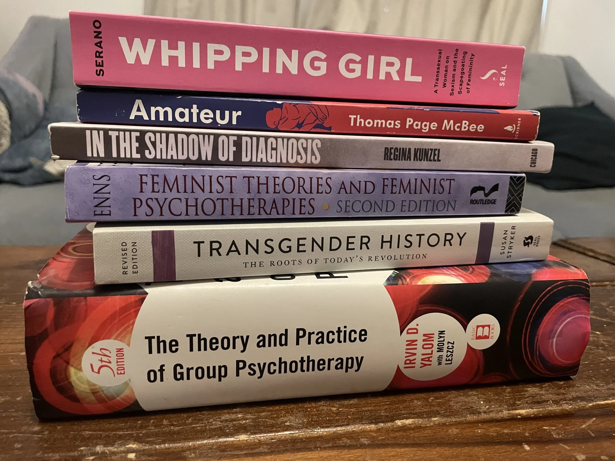 Stack of textbooks on psychology, gender studies, and psychotherapy with titles including 'Whipping Girl', 'Amateur', 'In the Shadow of Diagnosis', 'Feminist Theories and Feminist Psychotherapies', 'Transgender History', and 'The Theory and Practice of Group Psychotherapy'.