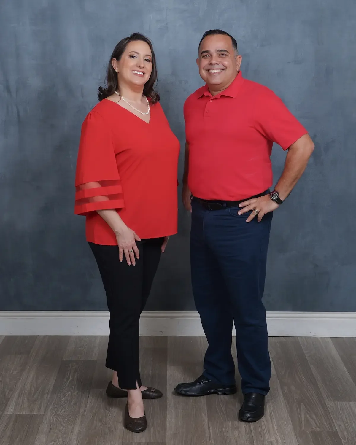 A man and woman standing together against a blue wall, both wearing red shirts and smiling.