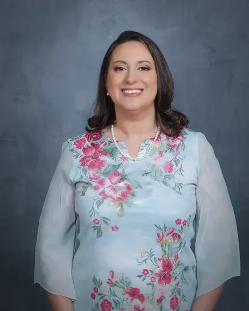 A woman smiling in a floral embroidered light blue dress with a pearl necklace.