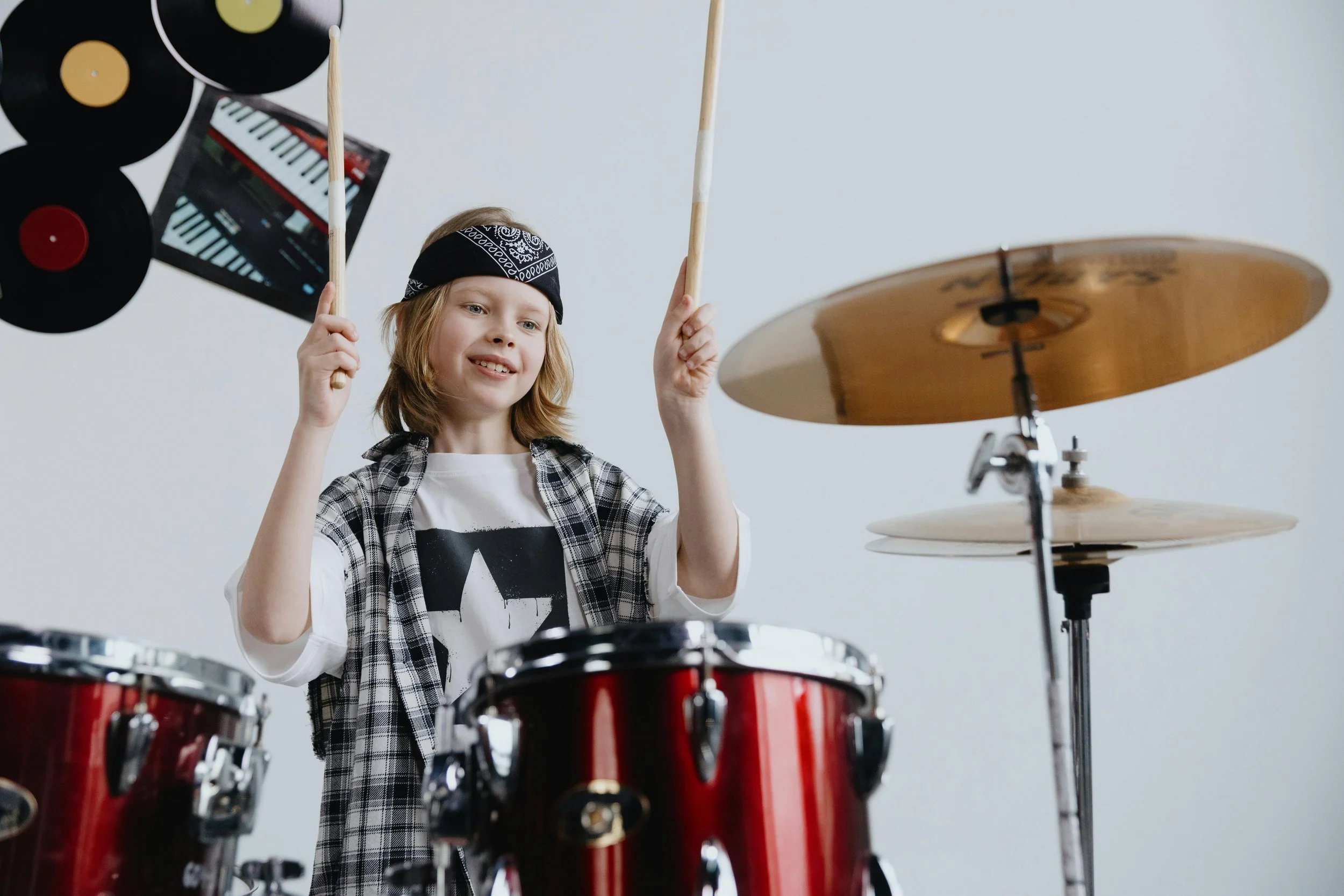 A young boy with long blond hair, sporting a blue bandana and a star-printed t-shirt, is playing a red drum kit. Both his sticks are raised up in the air.