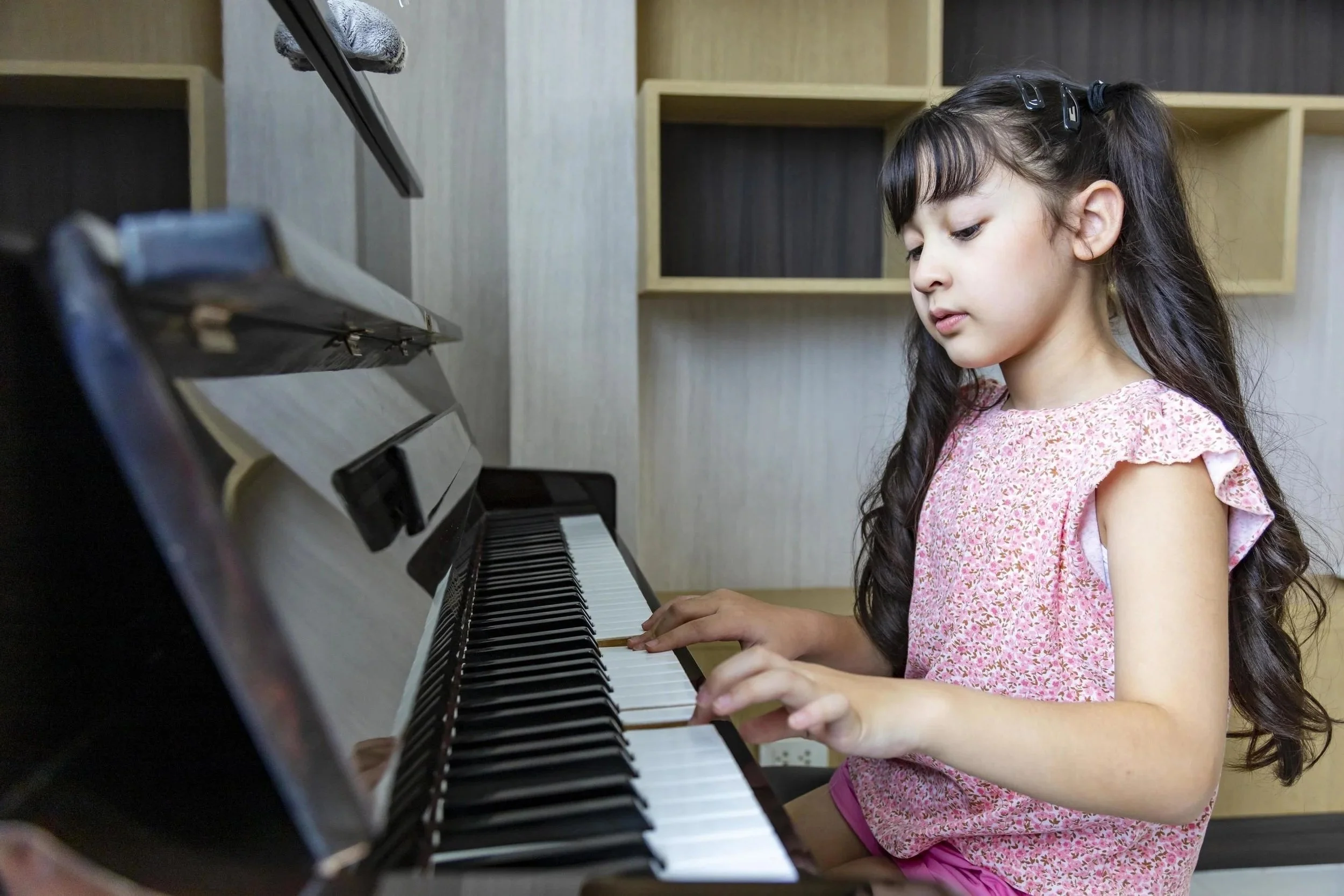 A young Asian girl with long brown hair, wearing a pink-speckled shirt, is practicing her upright piano at home.