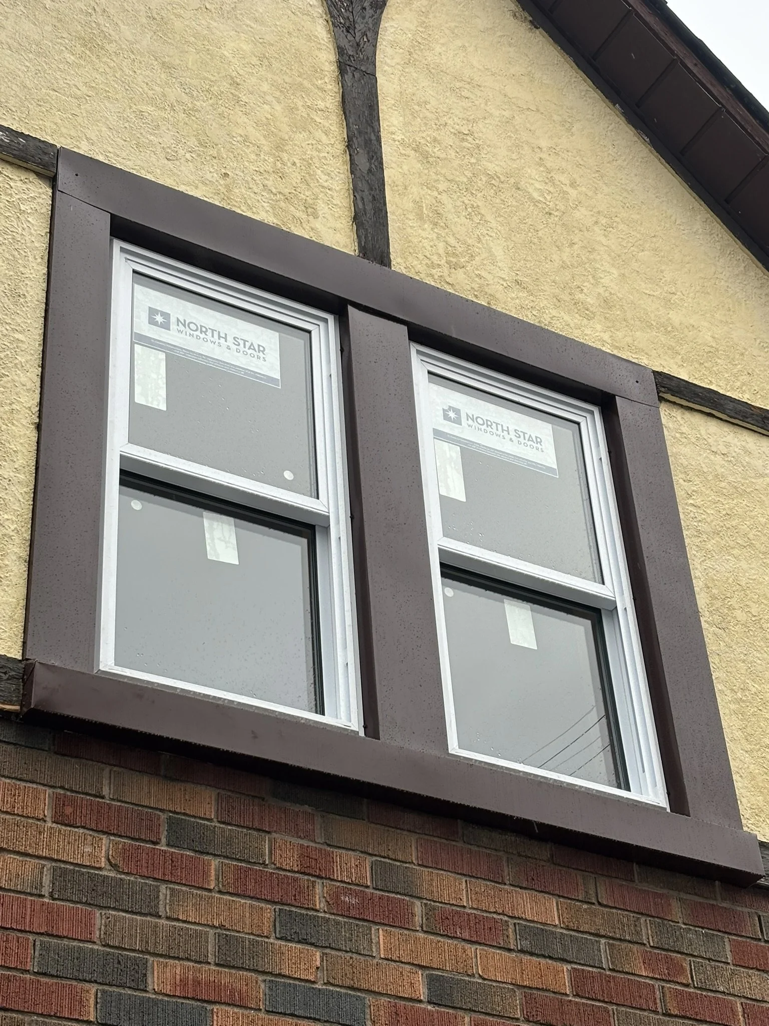 Double-hung window with brown trim on a yellow stucco house, with brick below and a dark roof overhang.