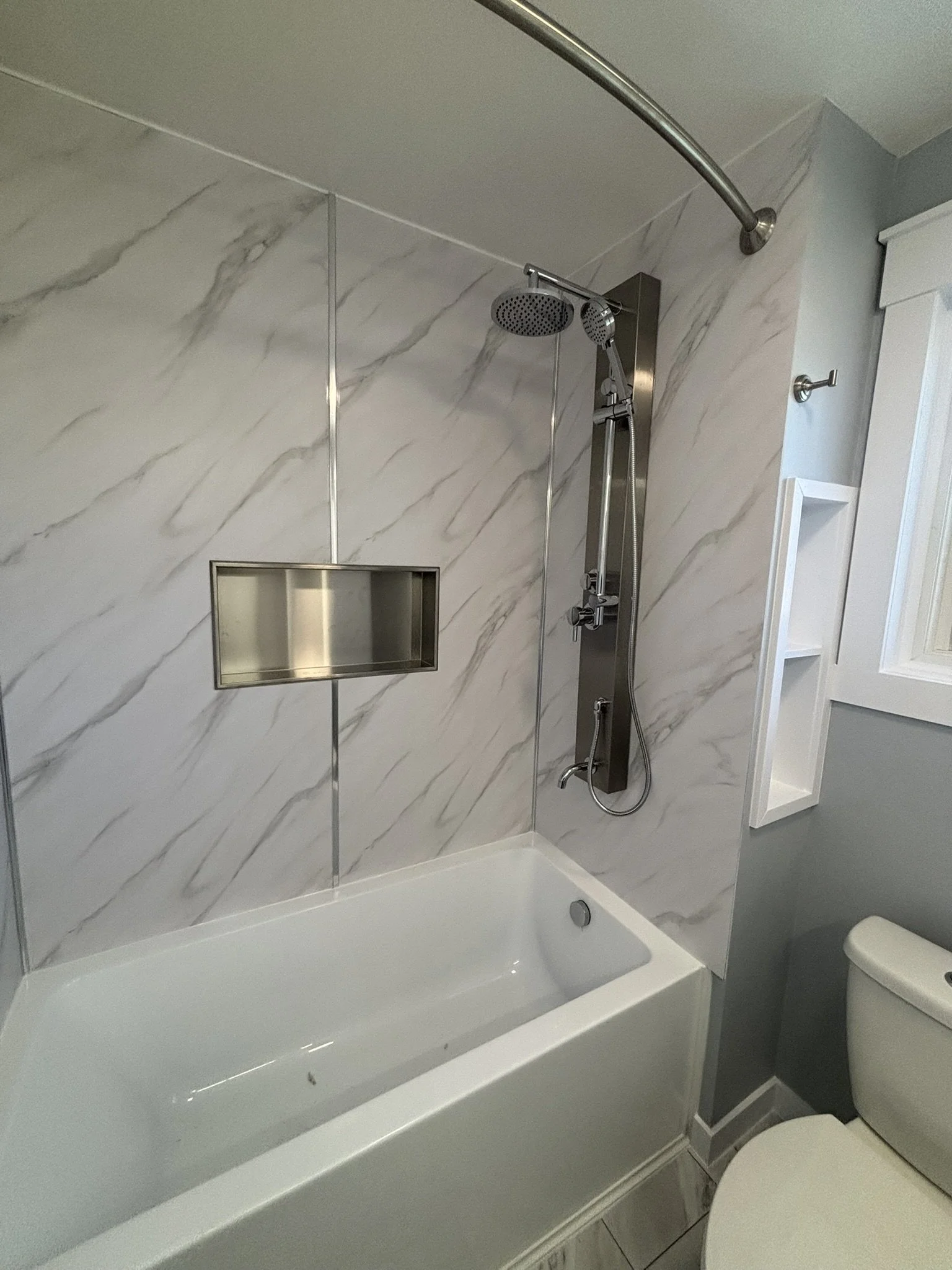 Bathroom shower with marble-patterned tile wall, stainless steel shower head and panel, built-in shower niche, and toilet beside the bathtub.