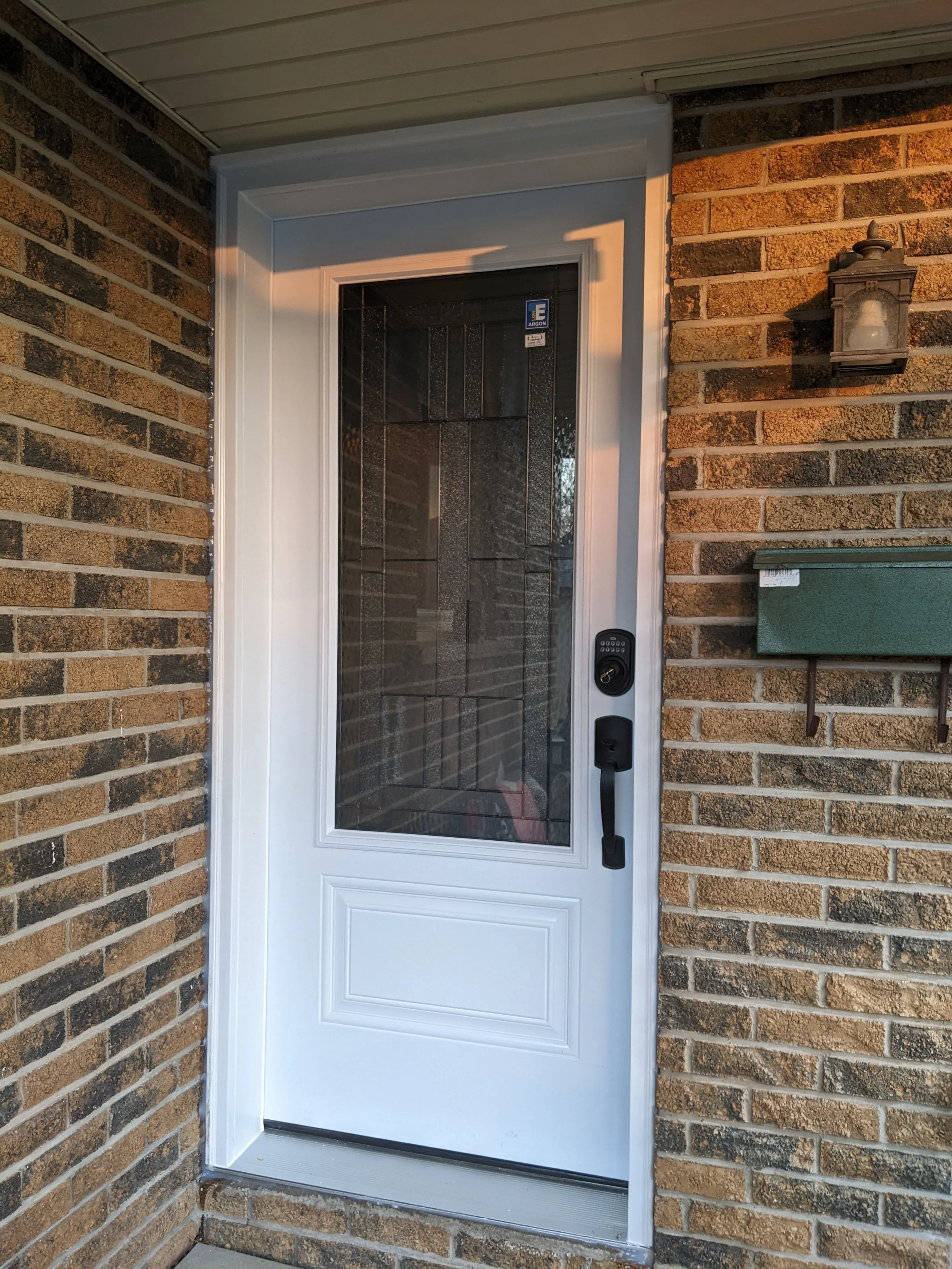 Front door of a house with brick siding, a white mesh storm door, a black electronic keypad lock, a mailbox, and a wall-mounted outdoor light fixture.