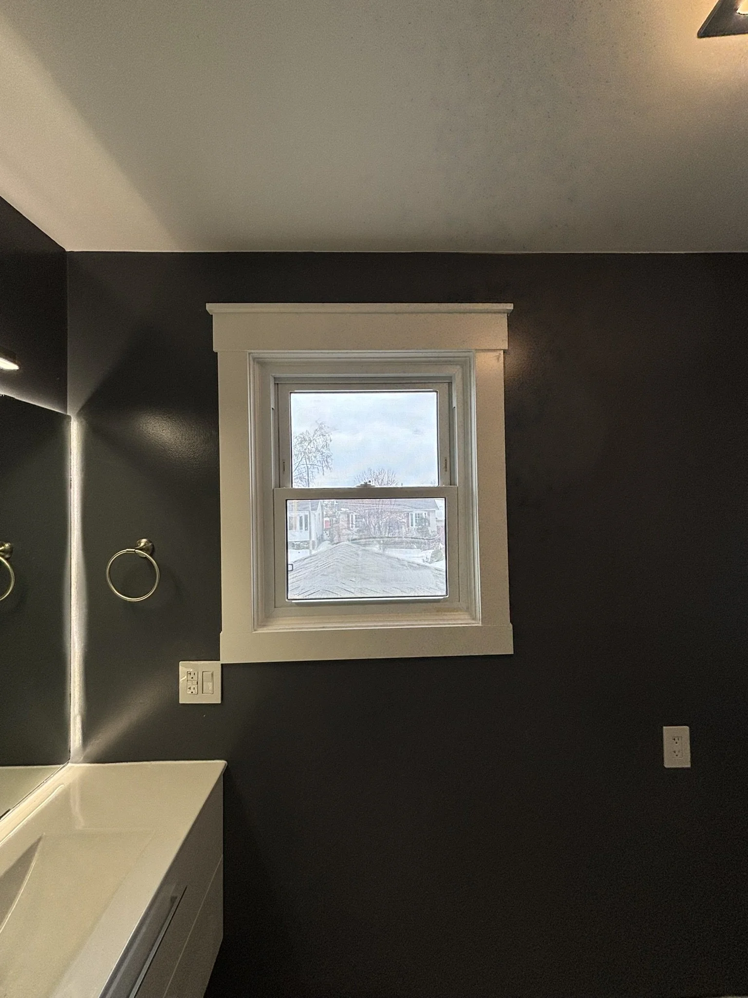 A bathroom window with white trim on a dark-colored wall, near a sink and mirror, with wall-mounted towel rings and electrical outlets.