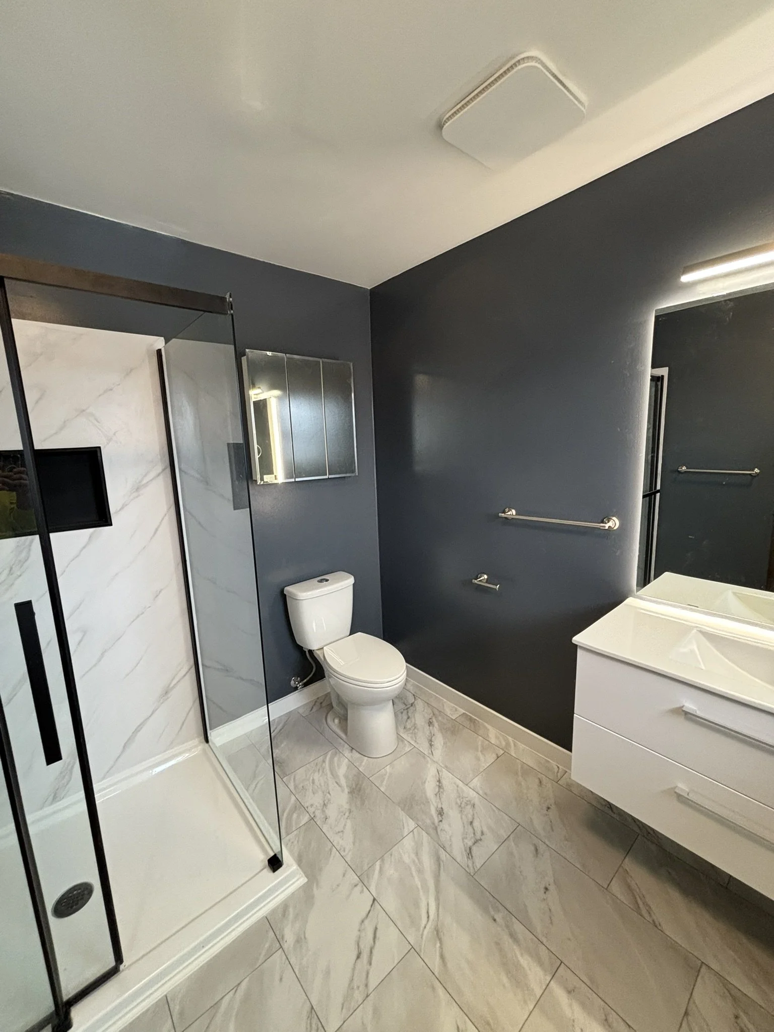 Modern bathroom with a glass shower, white toilet, and a white vanity with a mirror, dark blue walls, and marble tile flooring.