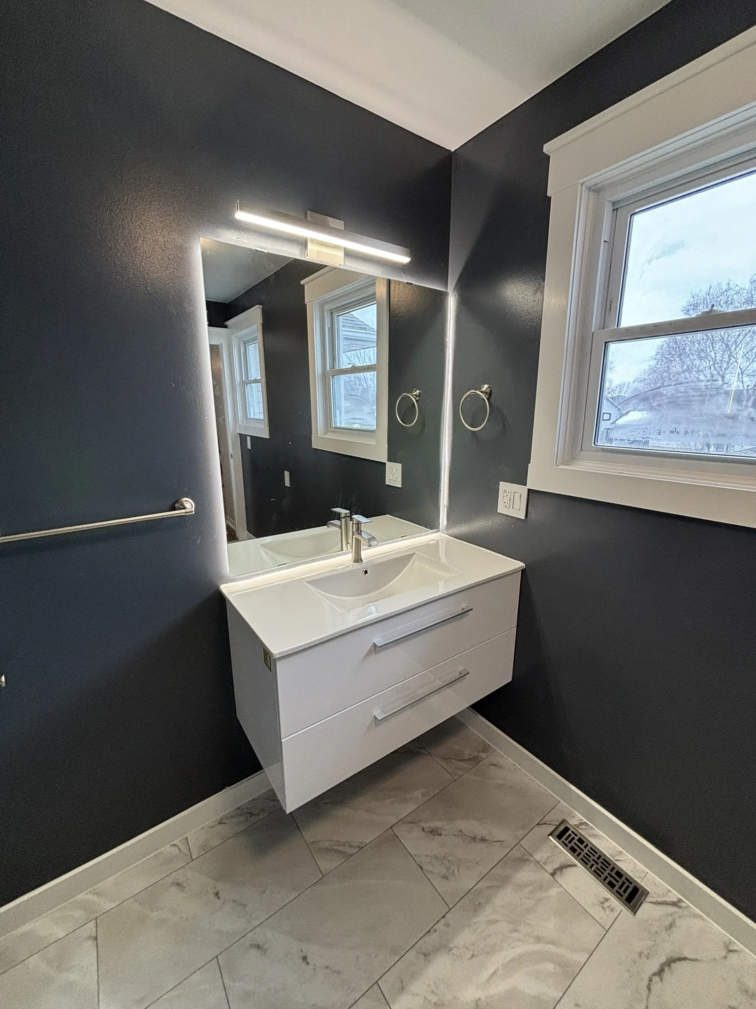 Modern bathroom with dark gray walls, white vanity with two drawers, a rectangular mirror, two towel rings, and a window showing a cloudy outdoor scene.