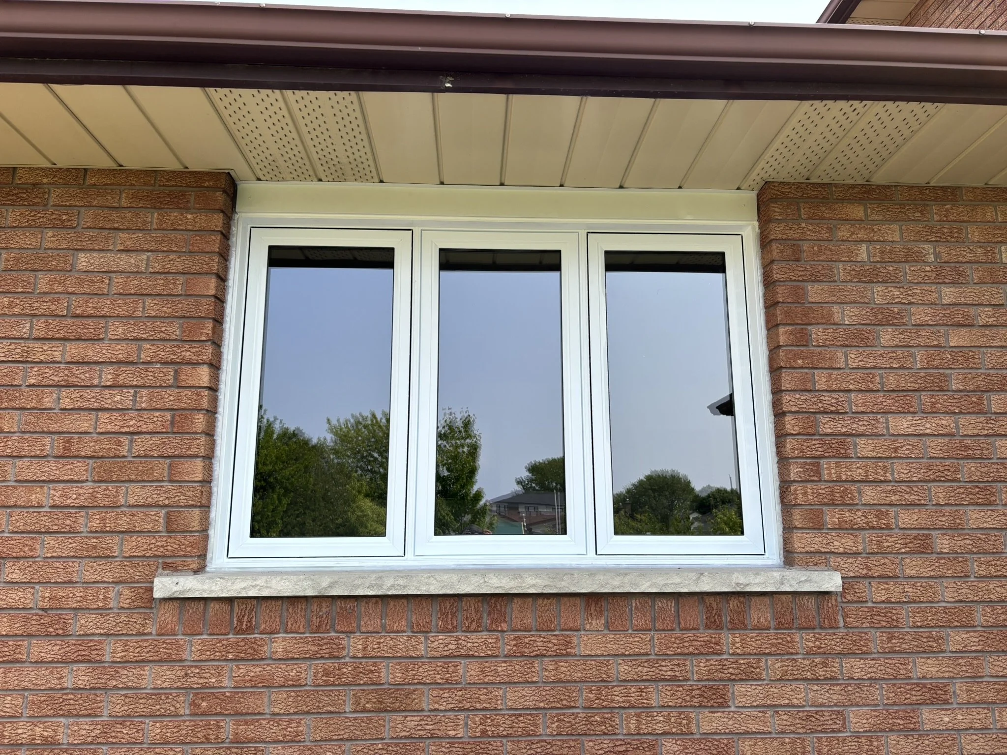 A large white-framed window with three sections, set into a red brick house wall, reflecting trees and the sky.