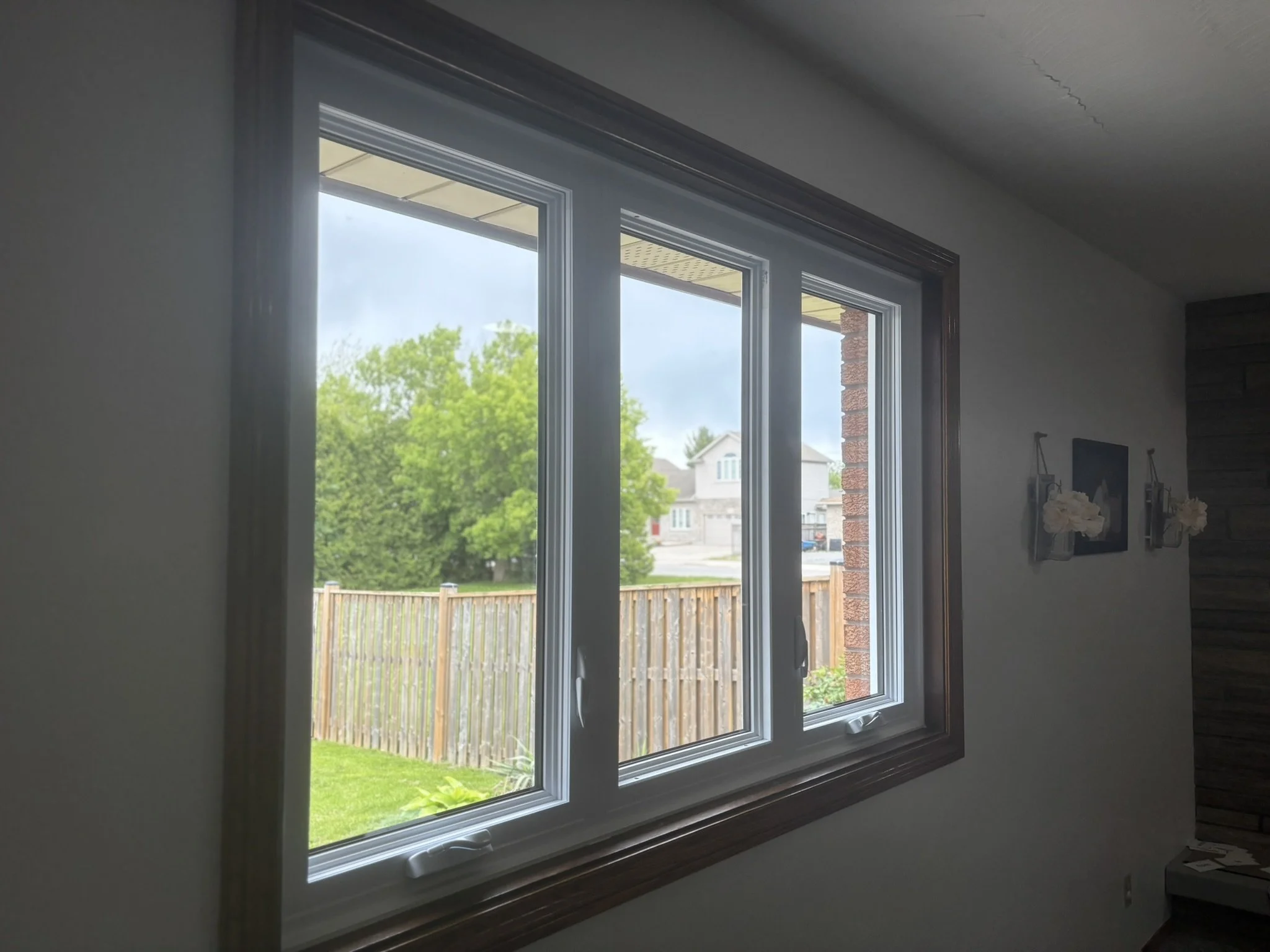 Indoor view of a double-pane window with a wooden frame, overlooking a backyard with a wooden fence, green grass, and neighboring houses under a cloudy sky.