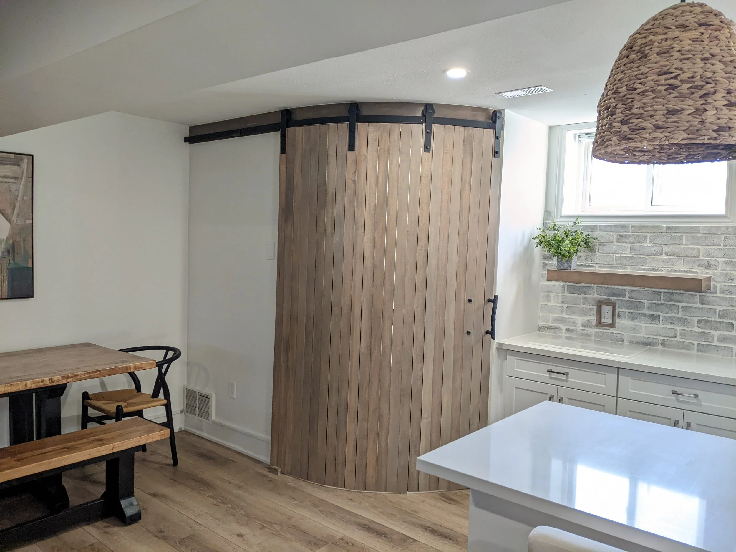 A corner of a modern kitchen with a white marble counter, a small potted plant on a brick wall shelf, a window, and a wooden sliding barn door on black metal hardware.