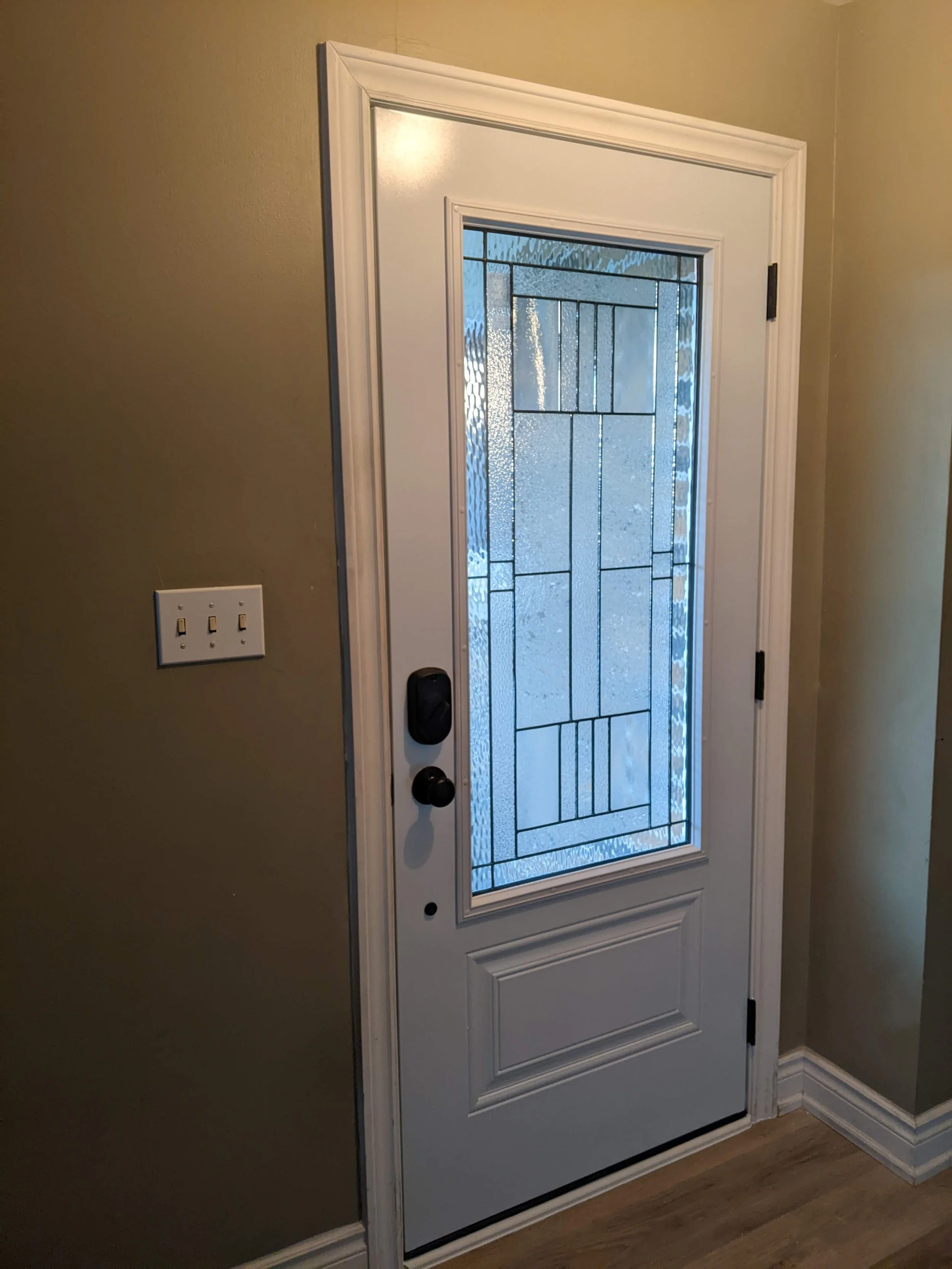 White front door with decorative glass panel, black handle and lock, surrounded by beige walls and light-colored wood flooring.