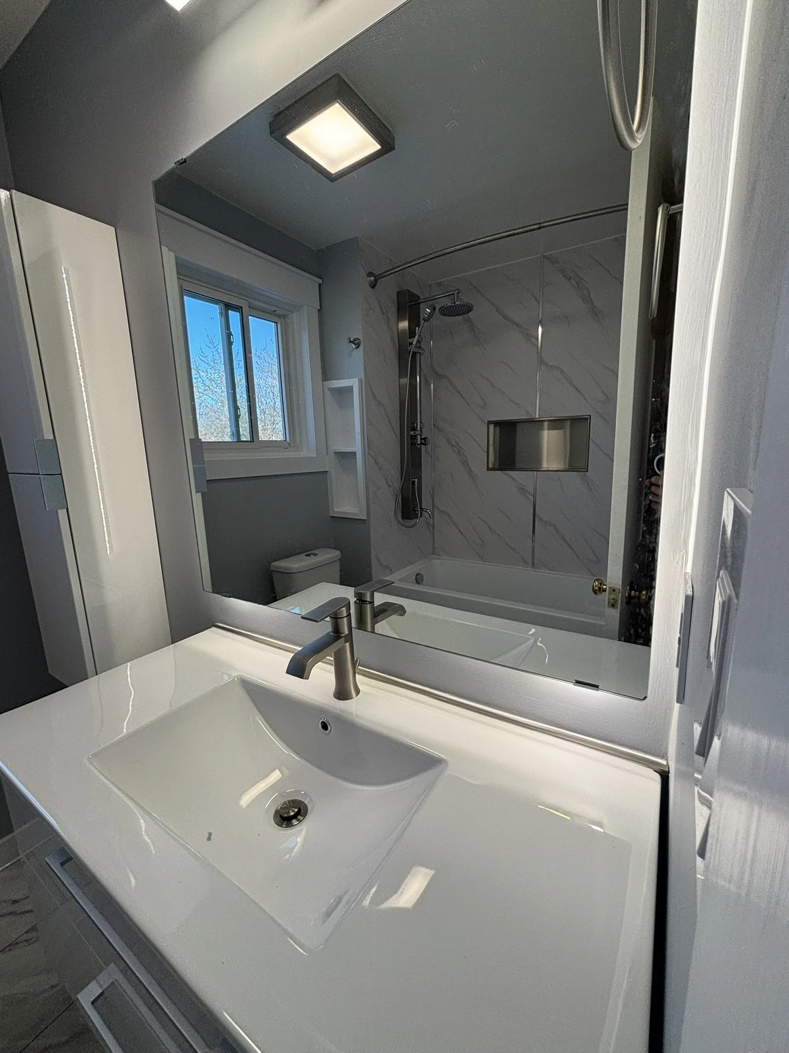 A modern bathroom with a white vanity sink, a large mirror, a window, and a bathtub with a shower. The walls are gray with marble accents, and there is a ceiling light fixture.