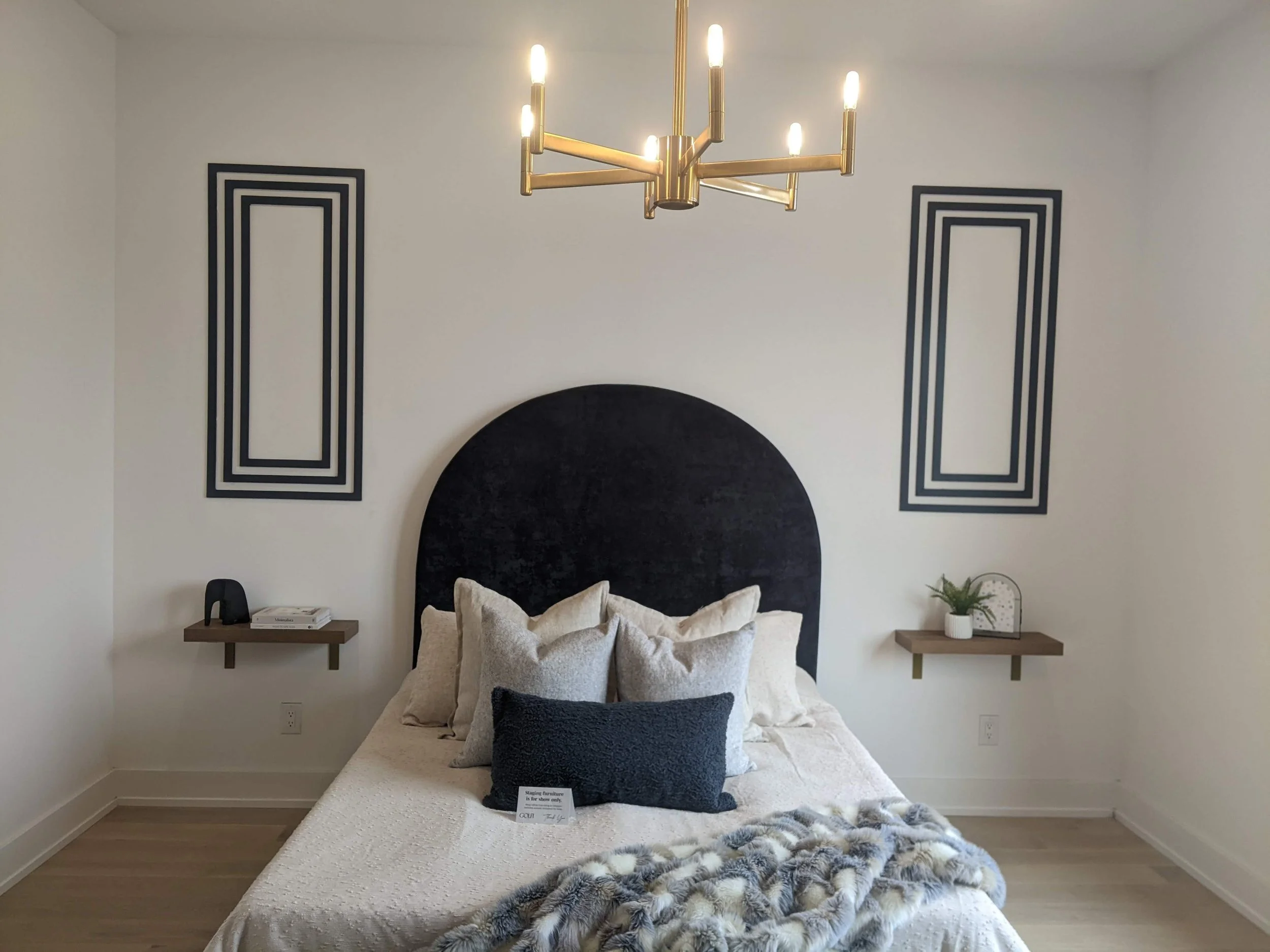 Modern bedroom with a black velvet headboard, beige bedding, and multiple pillows. Gold chandelier hanging from the ceiling, white walls with black rectangular wall art on either side of the bed, and floating wooden nightstands with decor items.