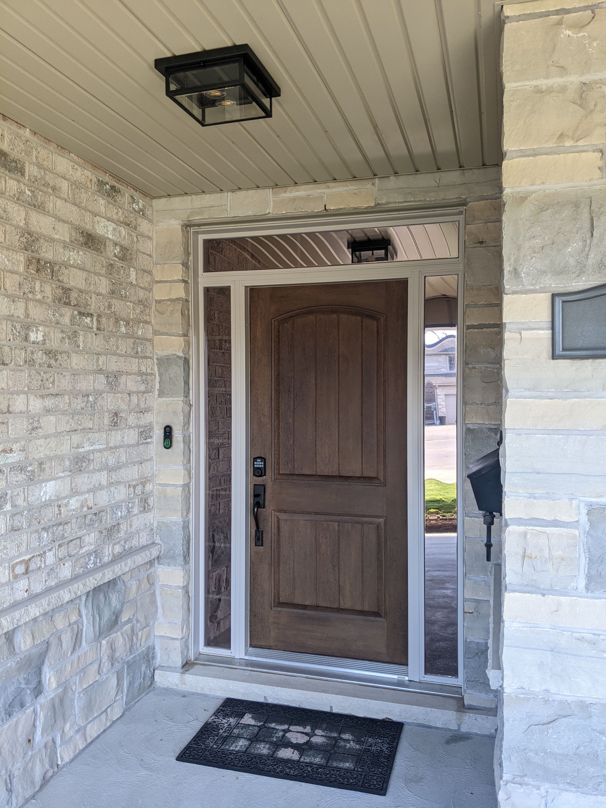 Front porch with a wooden door, security keypad, sliding screen door, decorative outdoor light, and a black doormat.