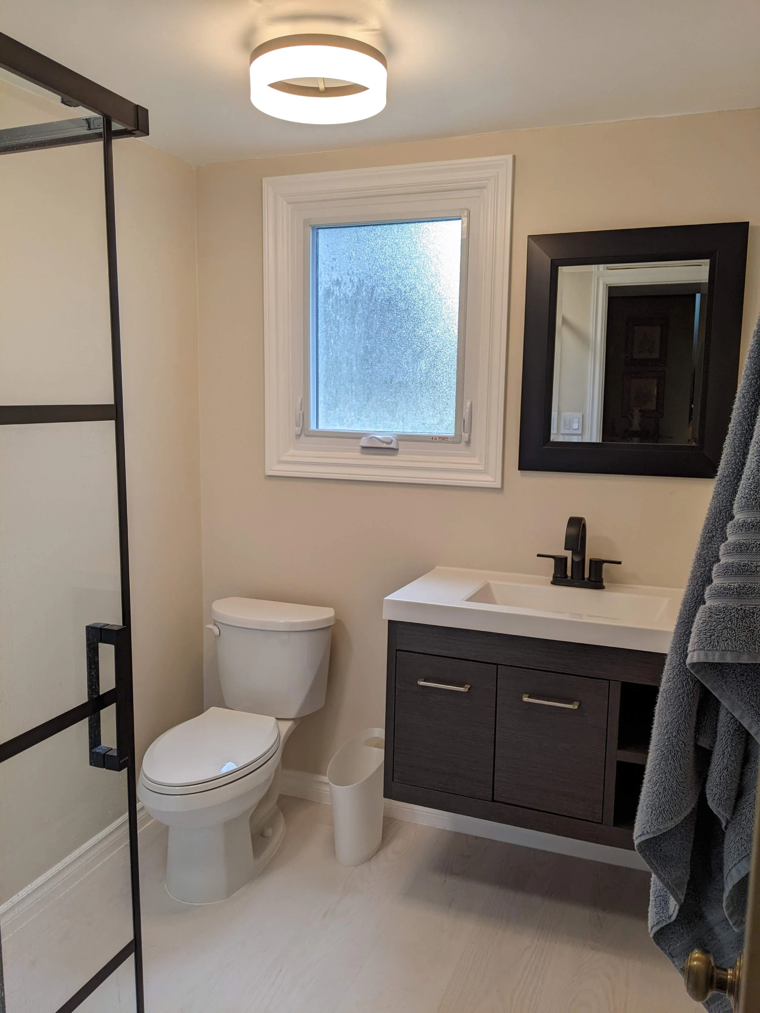 Bathroom with a toilet, a small vanity with a black faucet, a mirror, a frosted window, a gray towel, and a glass shower door.