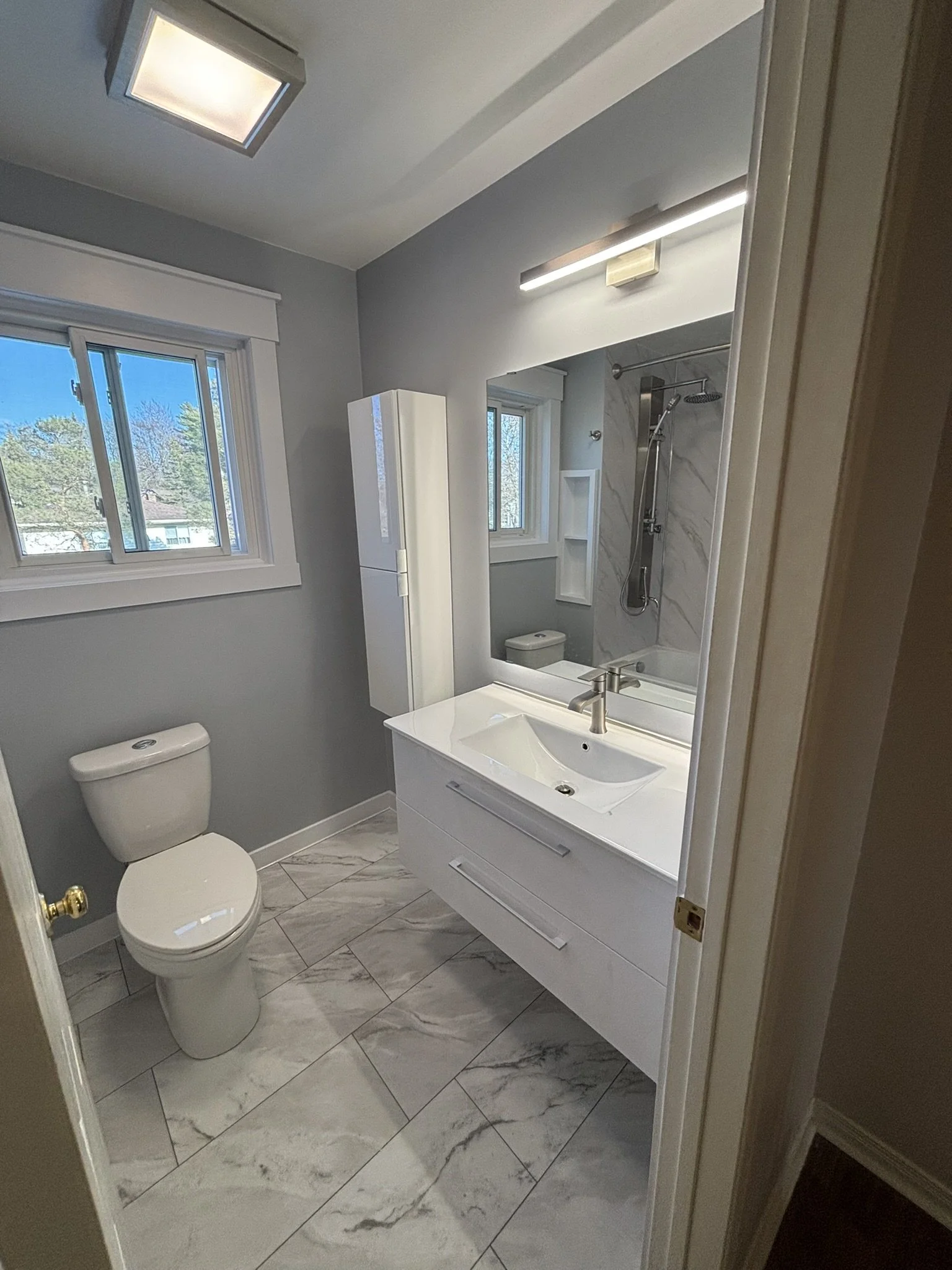 Modern bathroom with gray walls, white vanity with a rectangular sink, a large mirror, a shower with a glass door, a white toilet, and marble floor tiles.