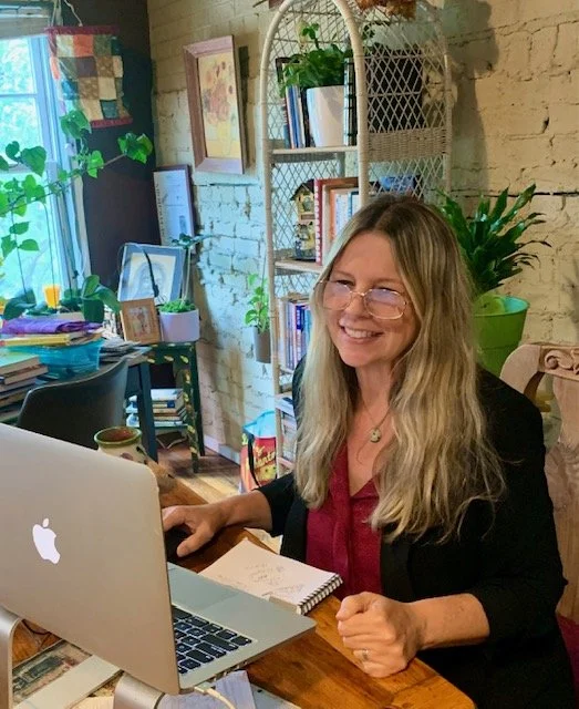 A woman with long blonde hair, glasses, and a black blazer sitting at a wooden desk, smiling, with a silver laptop, notebook, and pen in front of her in a cozy home office with plants, bookshelves, and wall decorations.