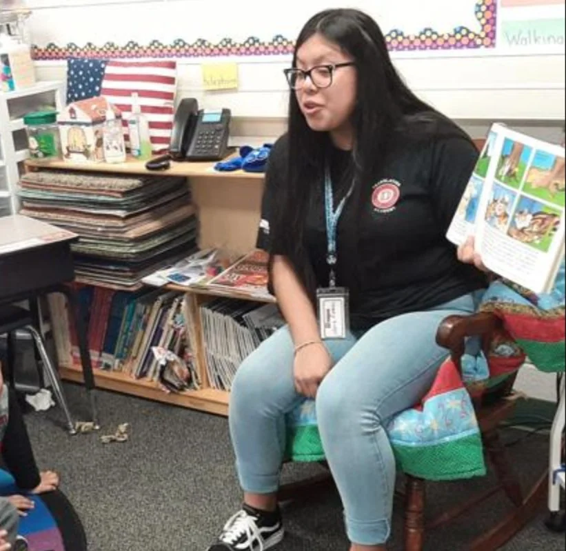 A woman with long black hair, glasses, and a black shirt sitting on a chair in a classroom, holding an open picture book, with books and school supplies around her.