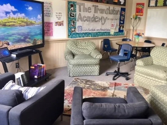 A classroom with a large screen displaying a nature scene, armchairs, a small table, and a corkboard with the sign 'The Traveling Academy' on the wall.