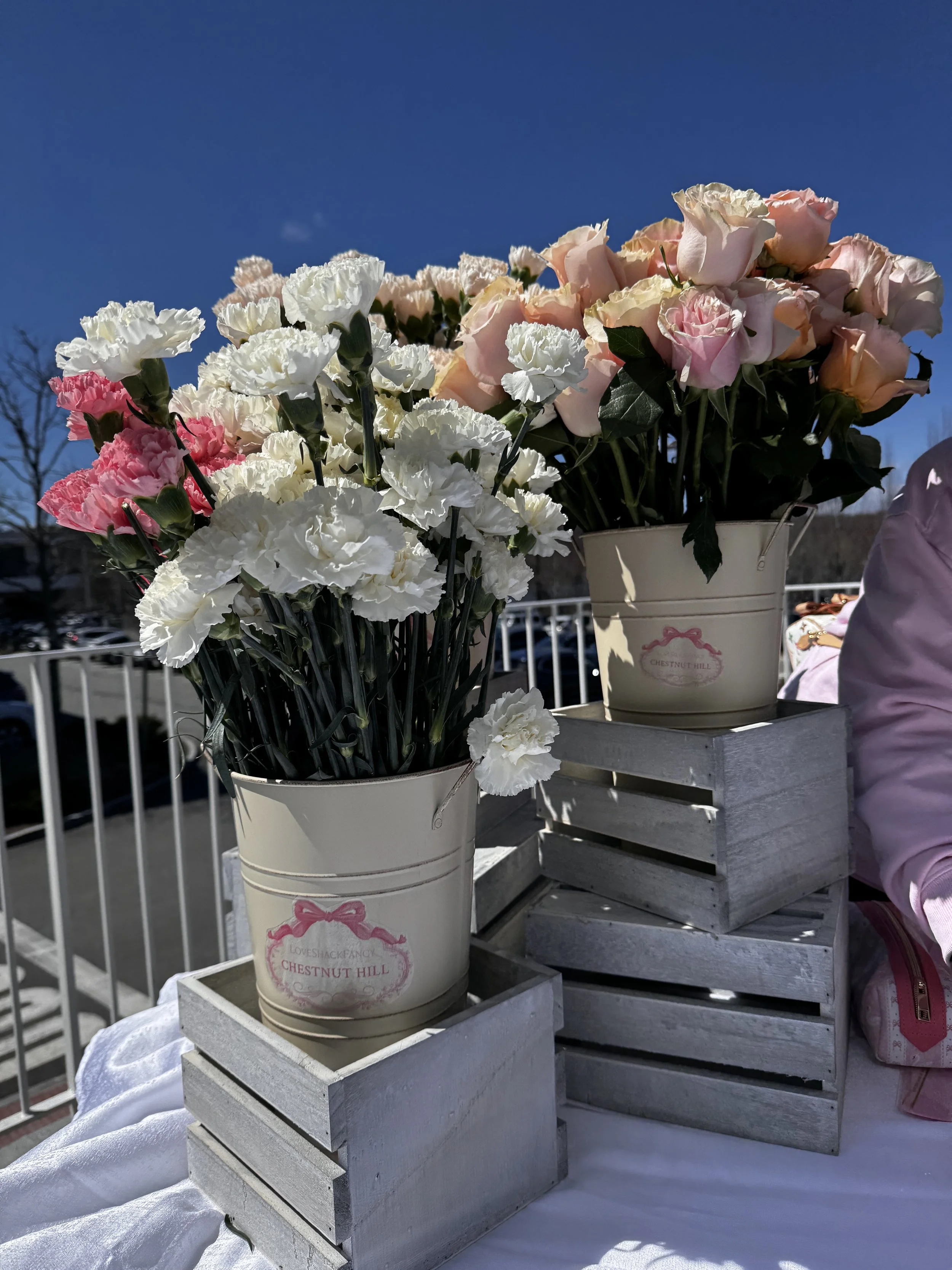 LoveShackFancy opening event with a flower bouquet bar set up with roses and carnations in white metal buckets adorned with custom brand stickers.