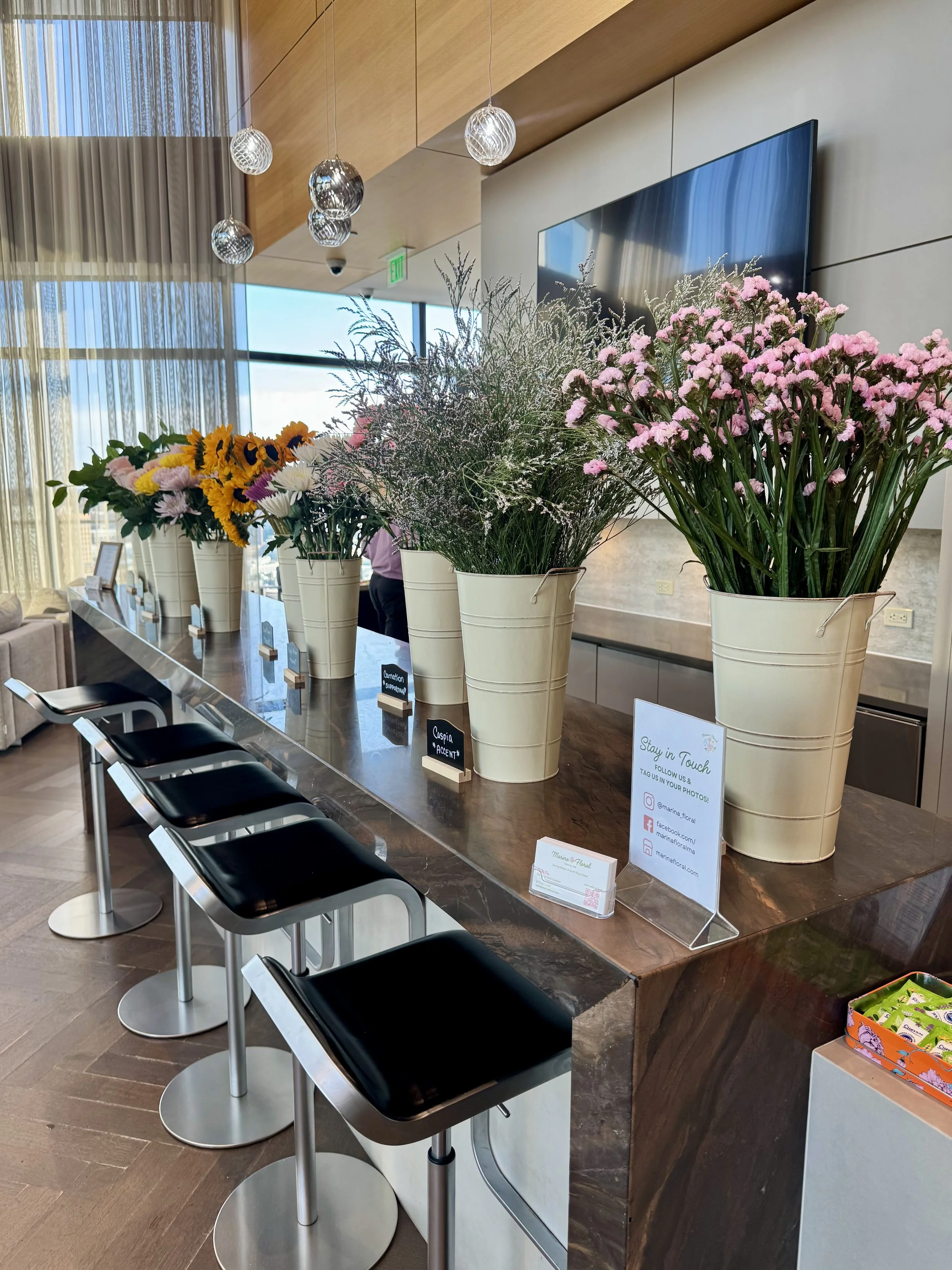 Indoor space with a long wooden counter and black stools. On the counter, there are several potted flower arrangements in white containers, with pink, yellow, and white flowers. In the background, there's a large flat screen mounted on the wall, a gl