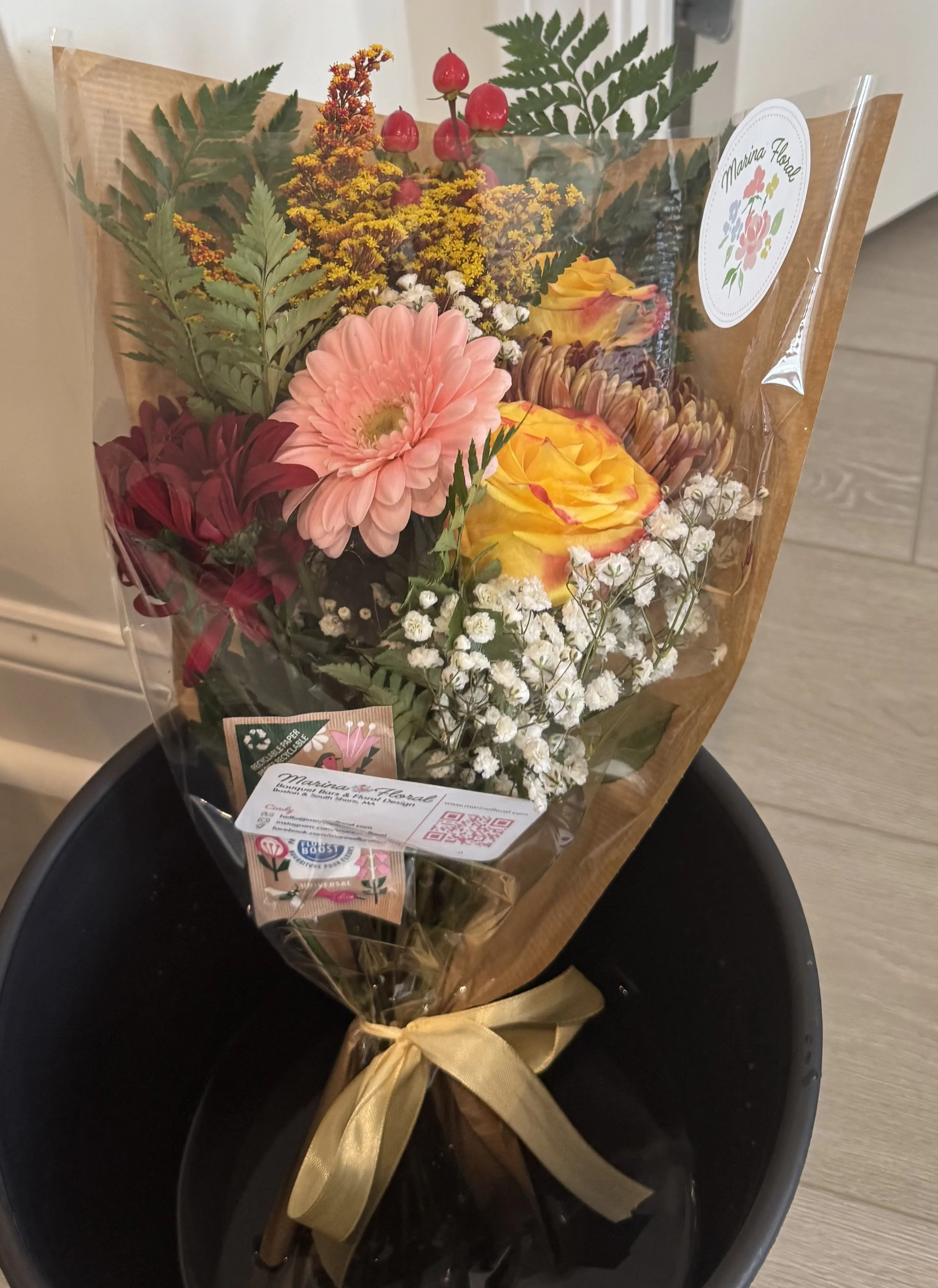 A bouquet of colorful flowers in a wrapped paper with a gold ribbon tied around the stems, placed on a black bucket.
