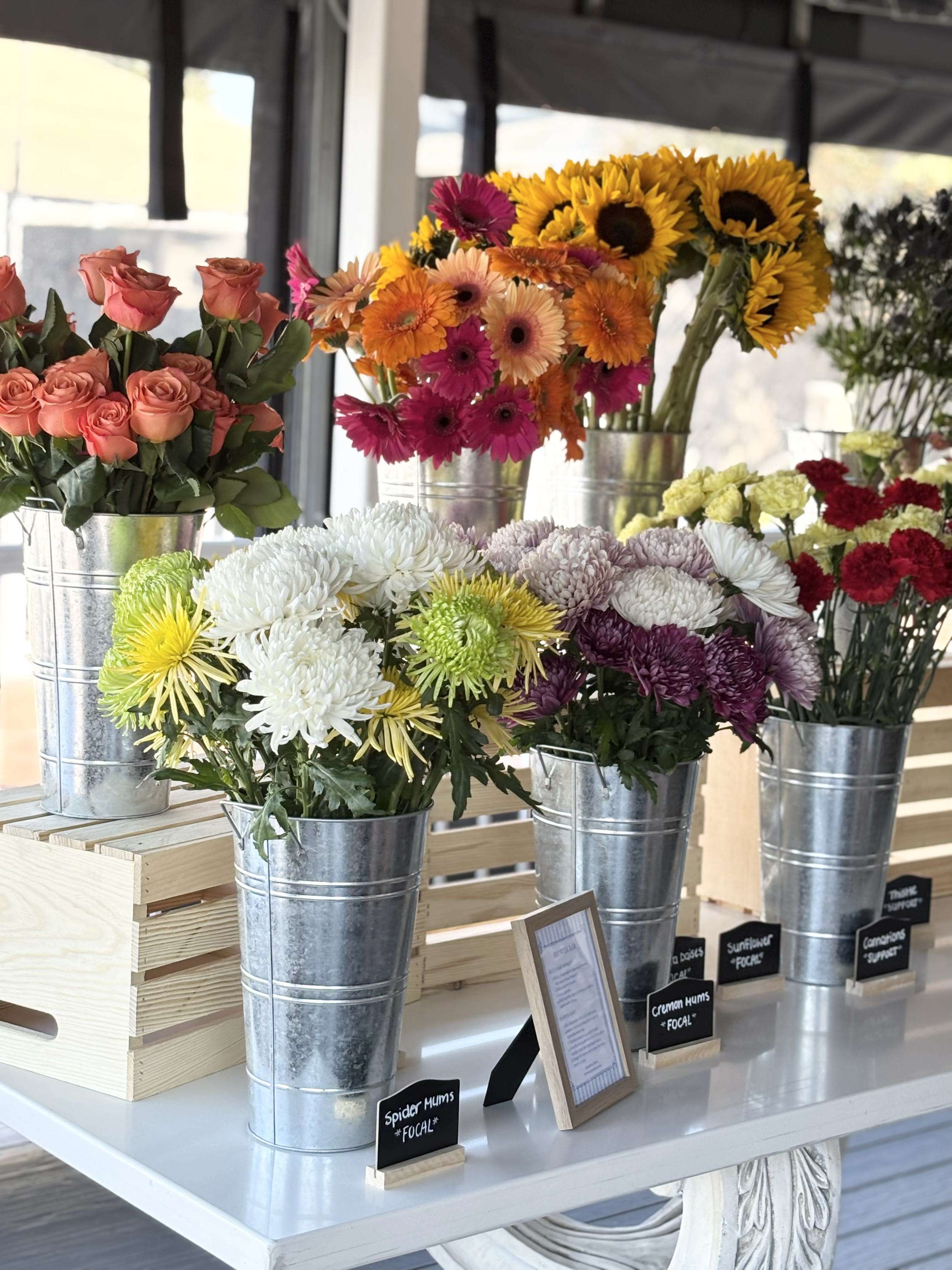 Various colorful flowers in metal buckets at a flower shop, including roses, sunflowers, daisies, mums, and carnations, with small black informational signs and a framed description on a white table.