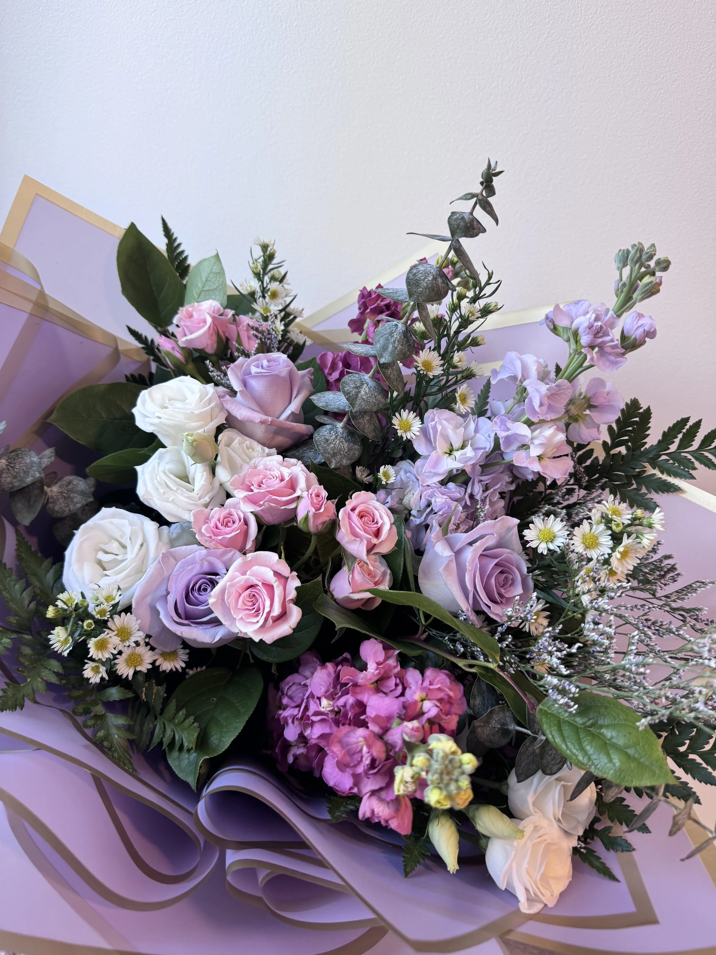 A bouquet of mixed flowers including pink and white roses, daisies, and greenery wrapped in purple and gold paper.
