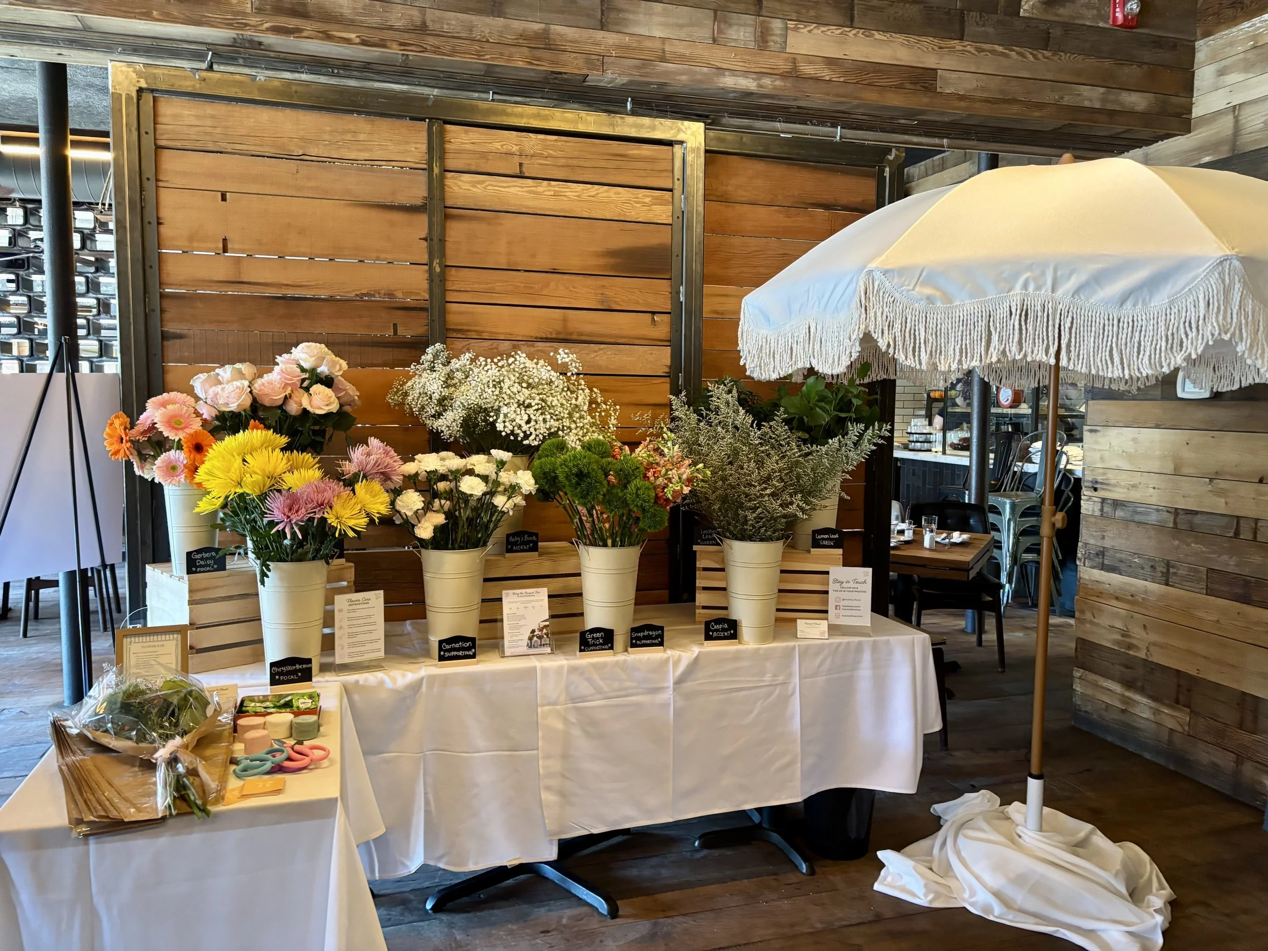 Display of various potted flowers on a table at an indoor market or shop. The background features a wooden panel wall, and there are signage labels beneath each plant for gardening-related purposes.