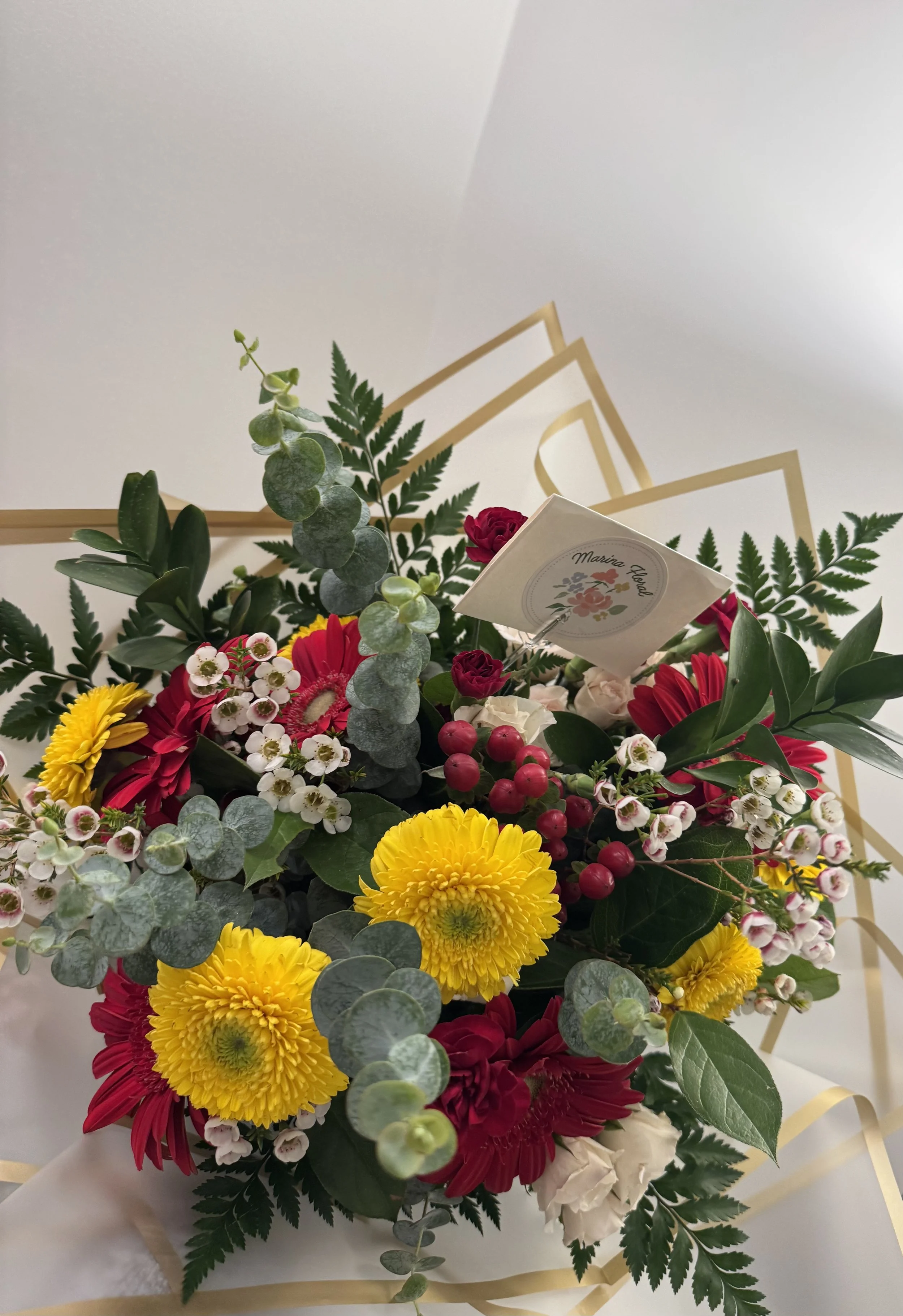 A colorful floral arrangement with yellow, red, white, and pink flowers, accented with green leaves and eucalyptus, tied with a cream-colored ribbon and a card that says 'Marina Flores,' against a white background.