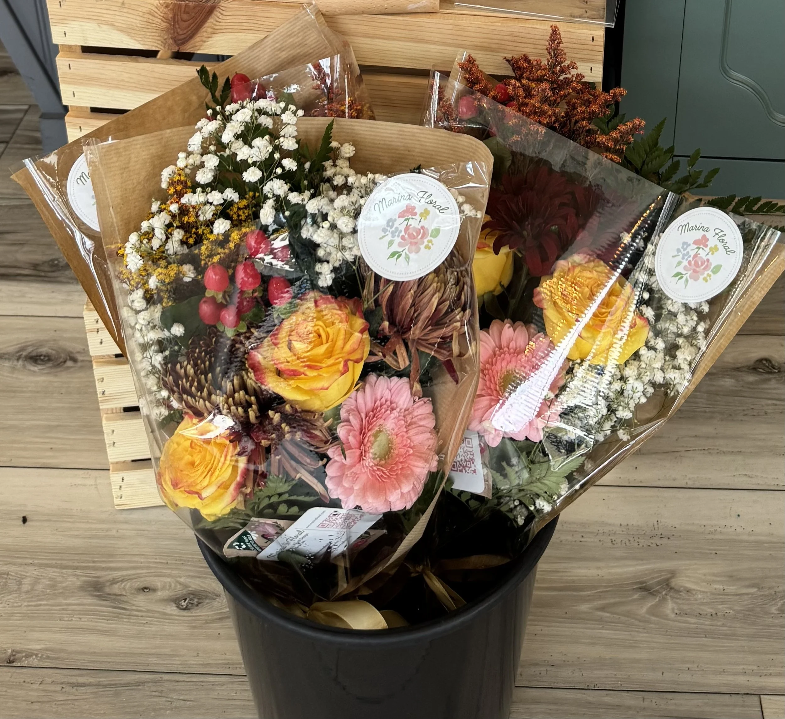 Assorted flower bouquets with pink, yellow, red, and white flowers wrapped in clear plastic, placed in a black bucket, on a wooden surface.