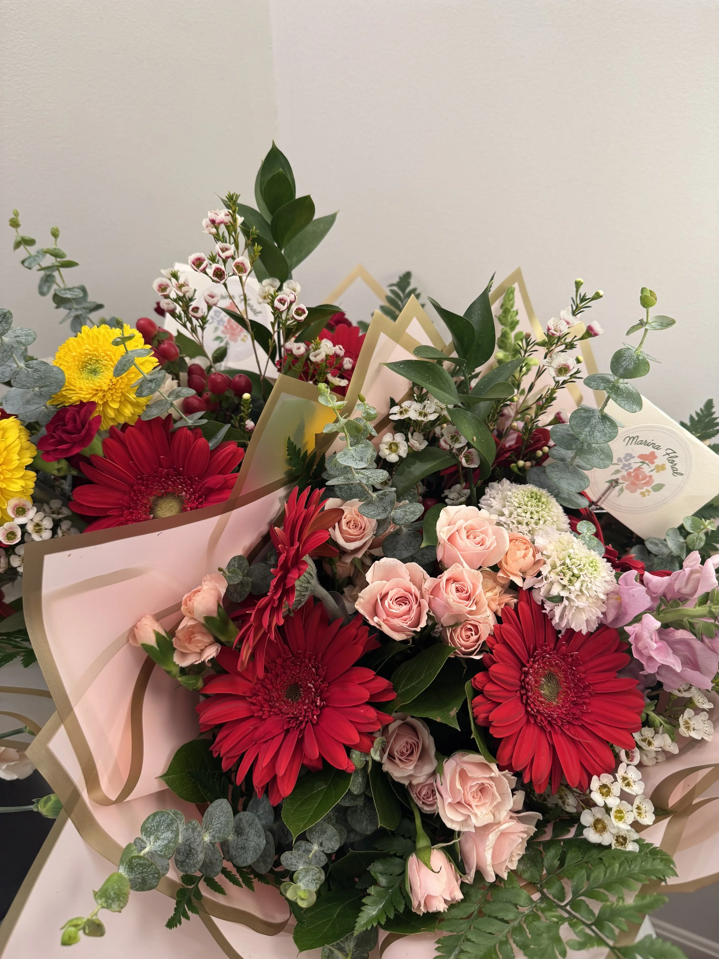 A bouquet of colorful flowers including red and yellow gerbera daisies, pink roses, small white flowers, and green foliage, wrapped in pink paper.
