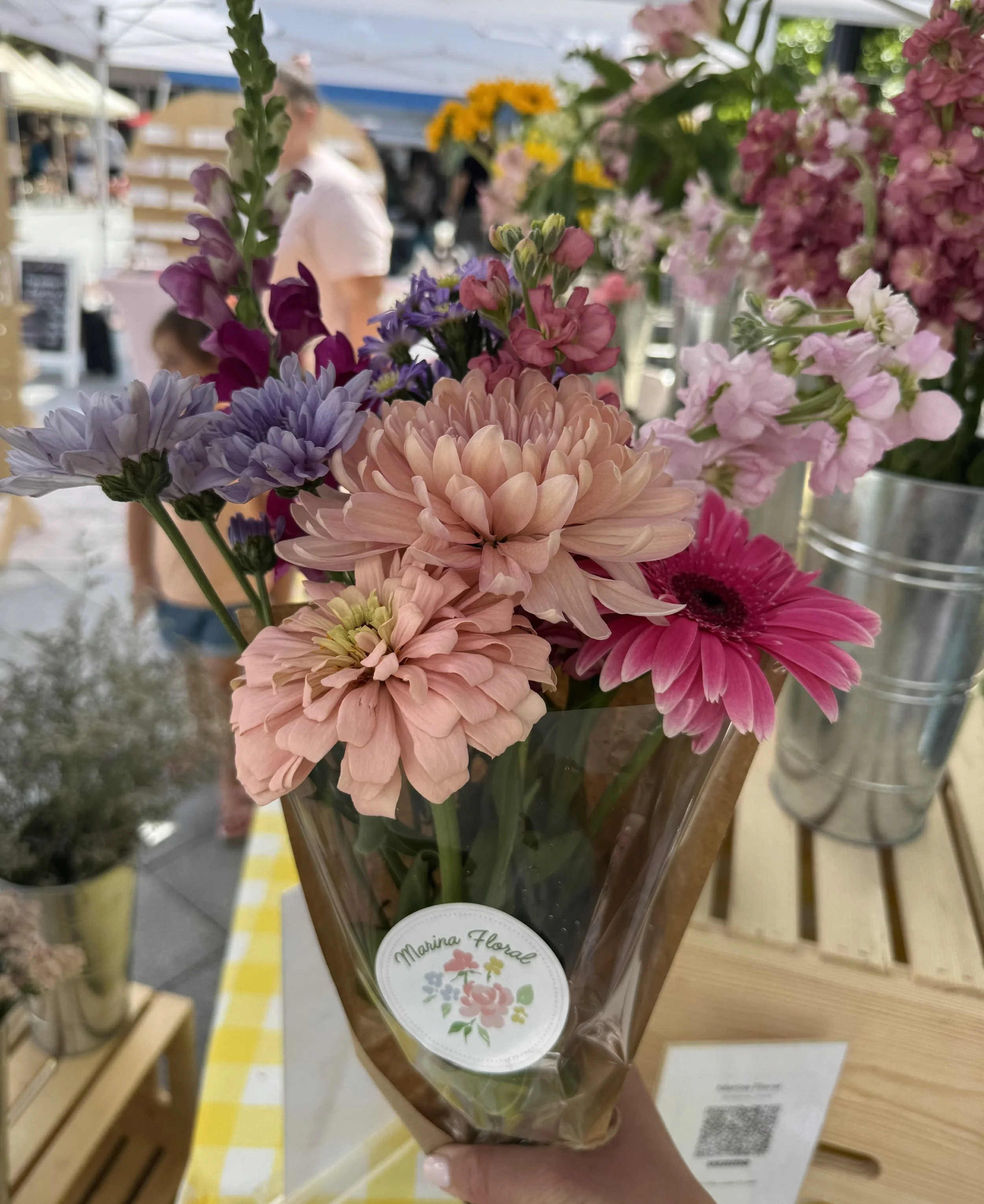 Bouquet of pink, purple, and yellow flowers in a clear plastic wrapping at a market stall. Floral Flower Bouquet Bar.