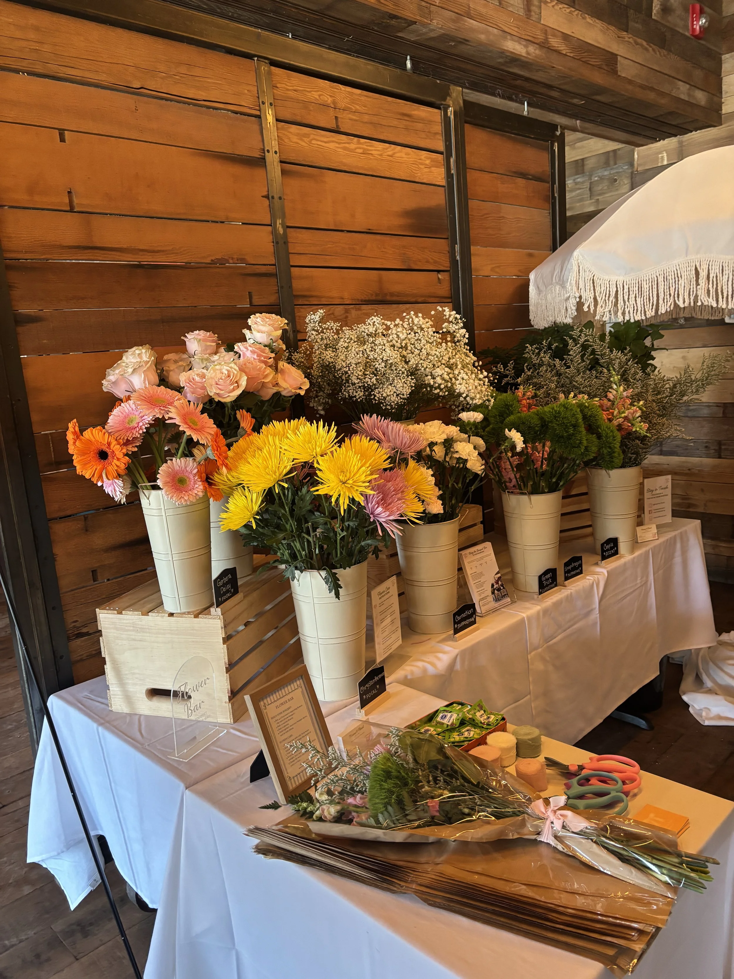A display of various flowers in white buckets at a flower shop, with some green foliage and packaging materials on a white tablecloth.