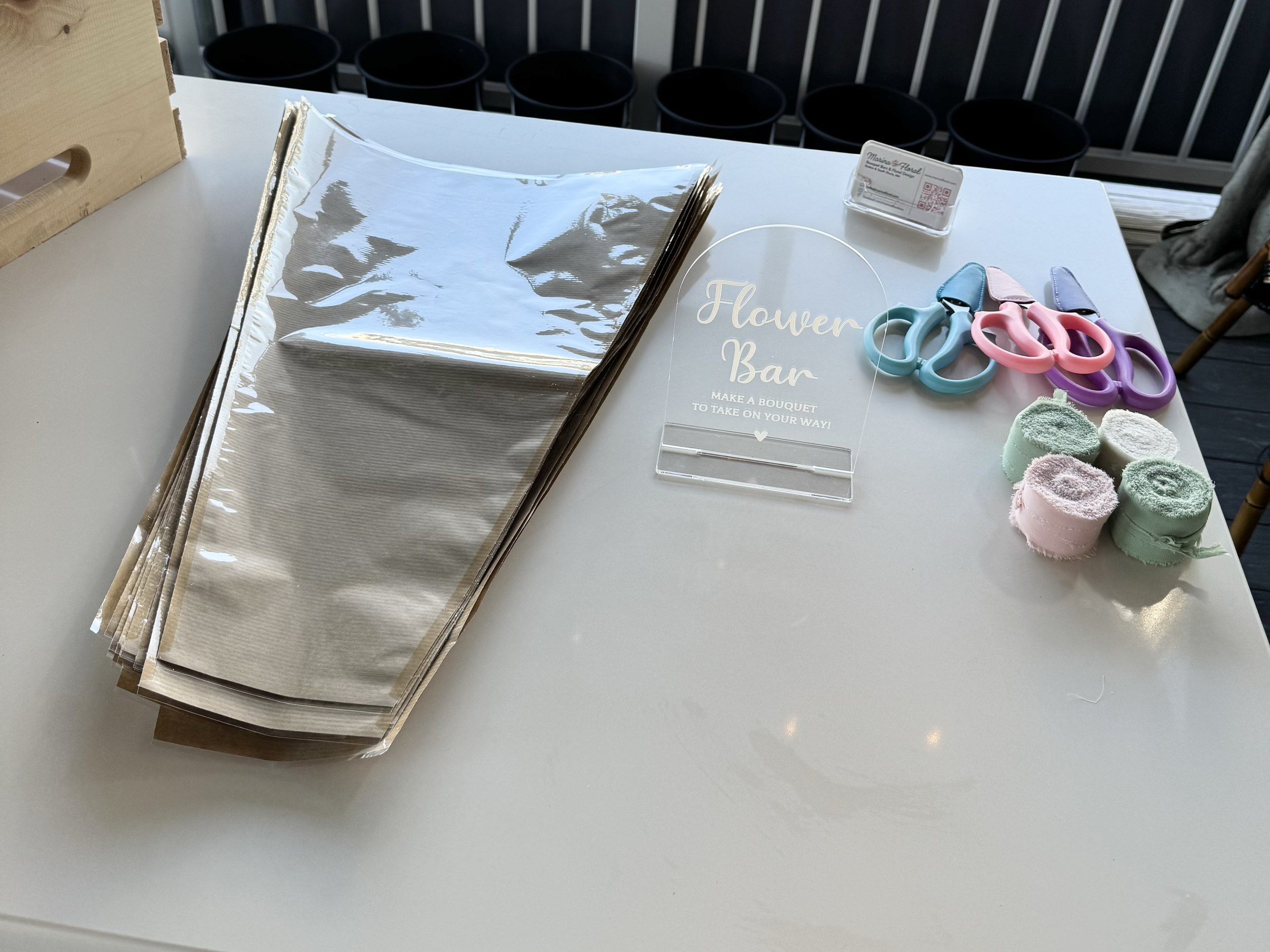 A white table displays a sign reading 'Flower Bar,' colored scissors, rolled pastel green, pink, and purple towels, a small clear sign holder with a QR code, and a stack of shiny metallic paper on the left side. Floral Flower Bouquet Bar.