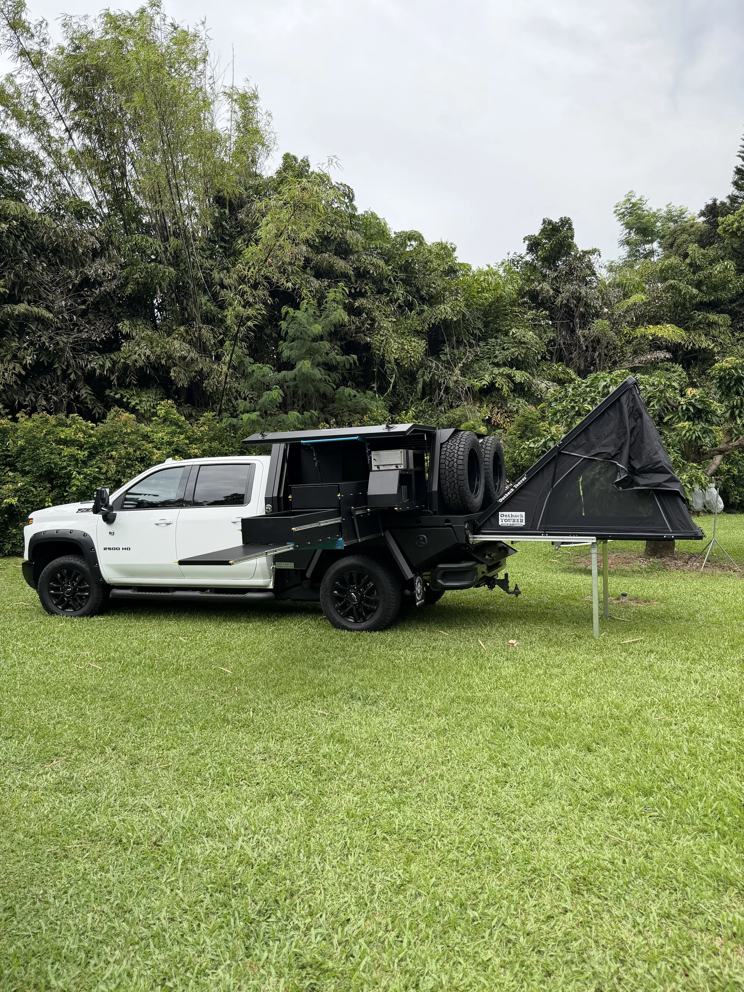White pickup truck with a black camping trailer attached, featuring a pop-up tent extended from the trailer on a grassy area with trees in the background.